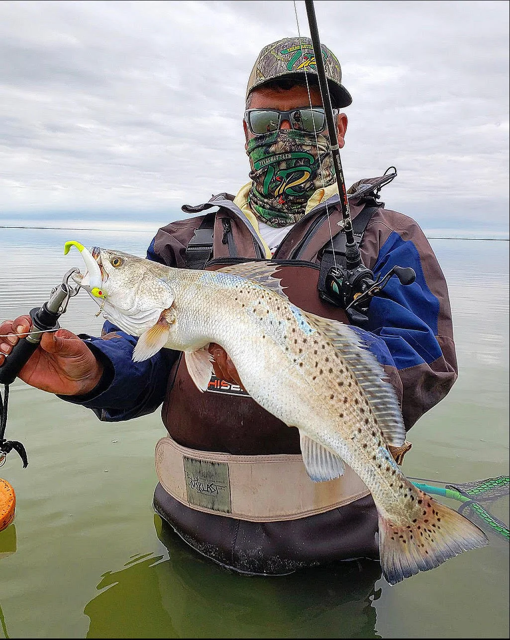 Person holding a speckled trout fish while standing in water, wearing fishing gear and a face mask.
