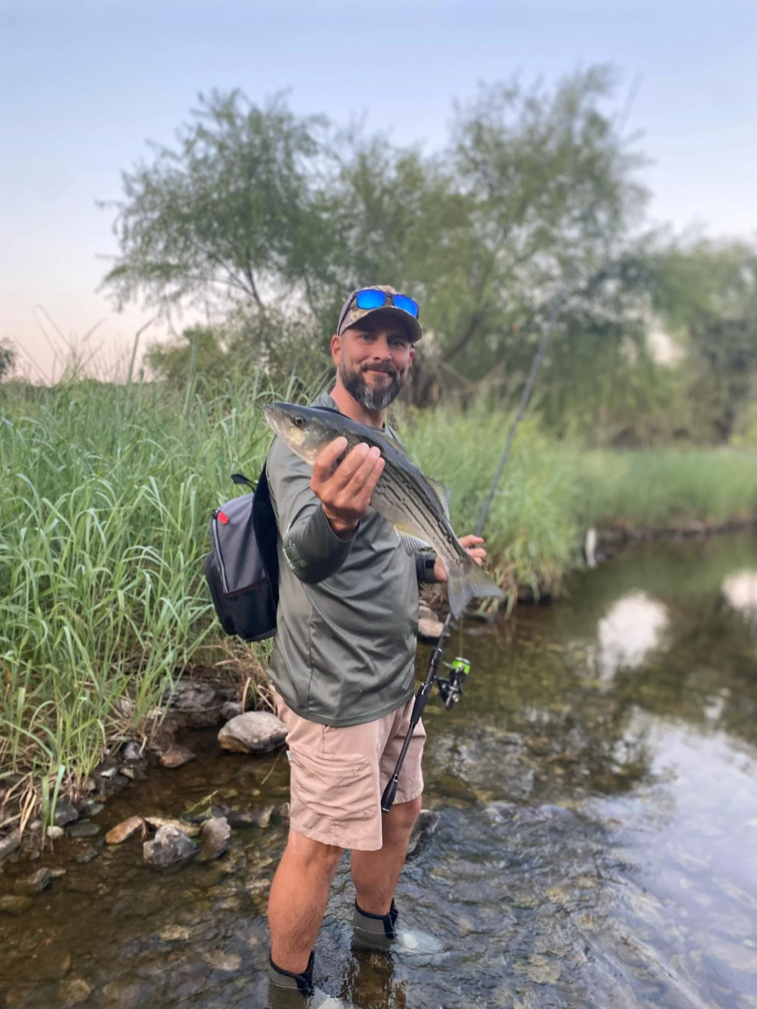 Man standing in shallow water holding a fish with a fishing rod in a natural setting with trees and grass.