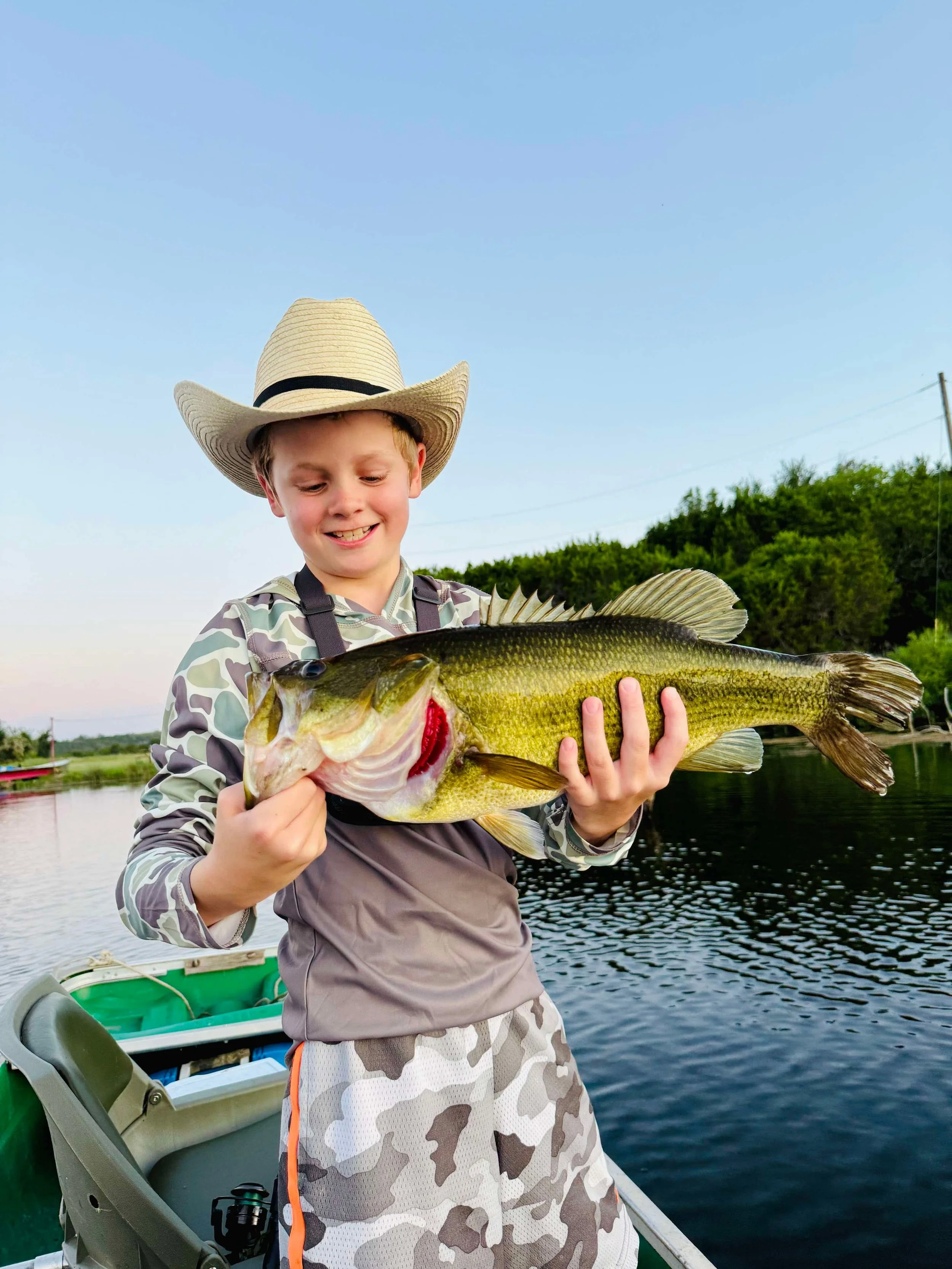 A young boy wearing a straw hat and camouflage clothing is smiling and holding a large fish he caught, standing on a boat near a lake with trees in the background during daylight.