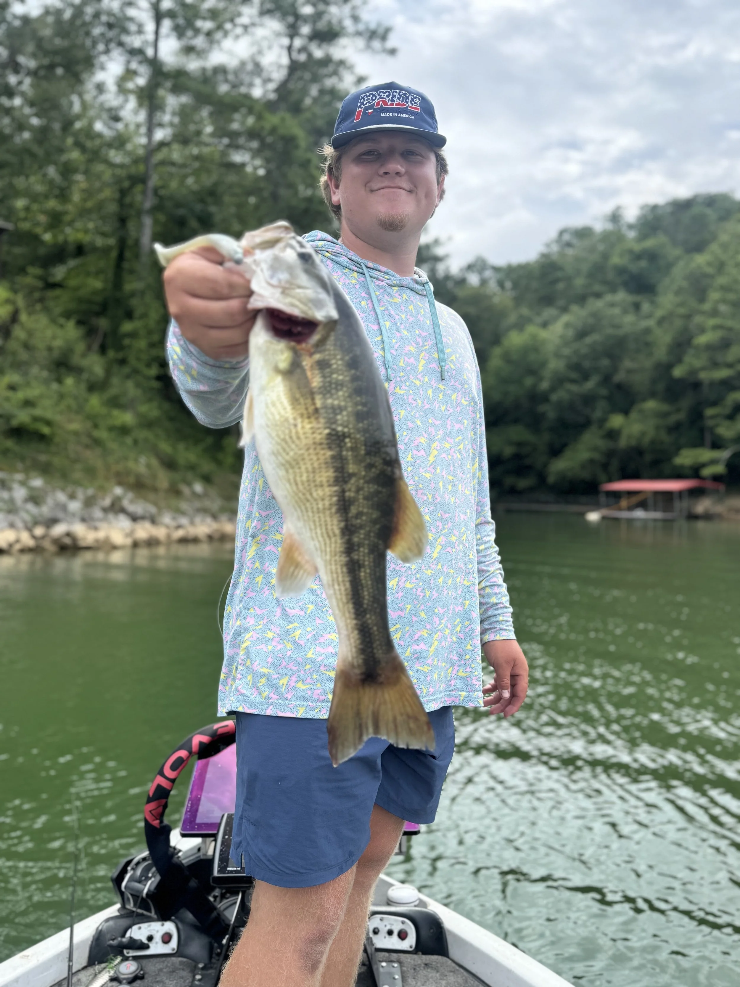 A young man in a colorful hoodie and blue shorts is standing on a boat holding a large fish, with a lake and green trees in the background.
