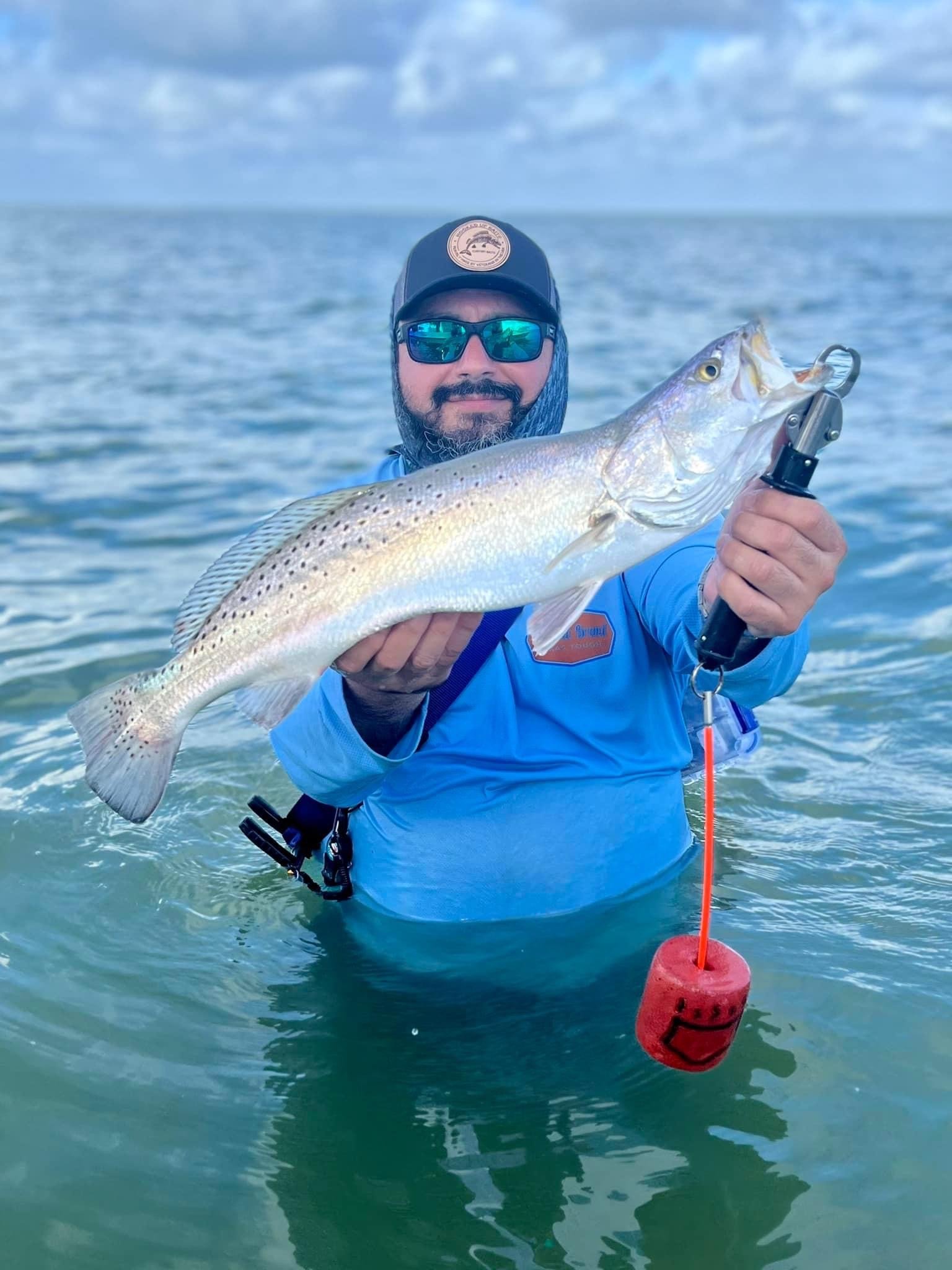 Person standing in water holding a large fish, wearing a blue shirt, sunglasses, cap, and neck gaiter. Fish is silver with spots, held with fish grip tool. Ocean and sky in background.