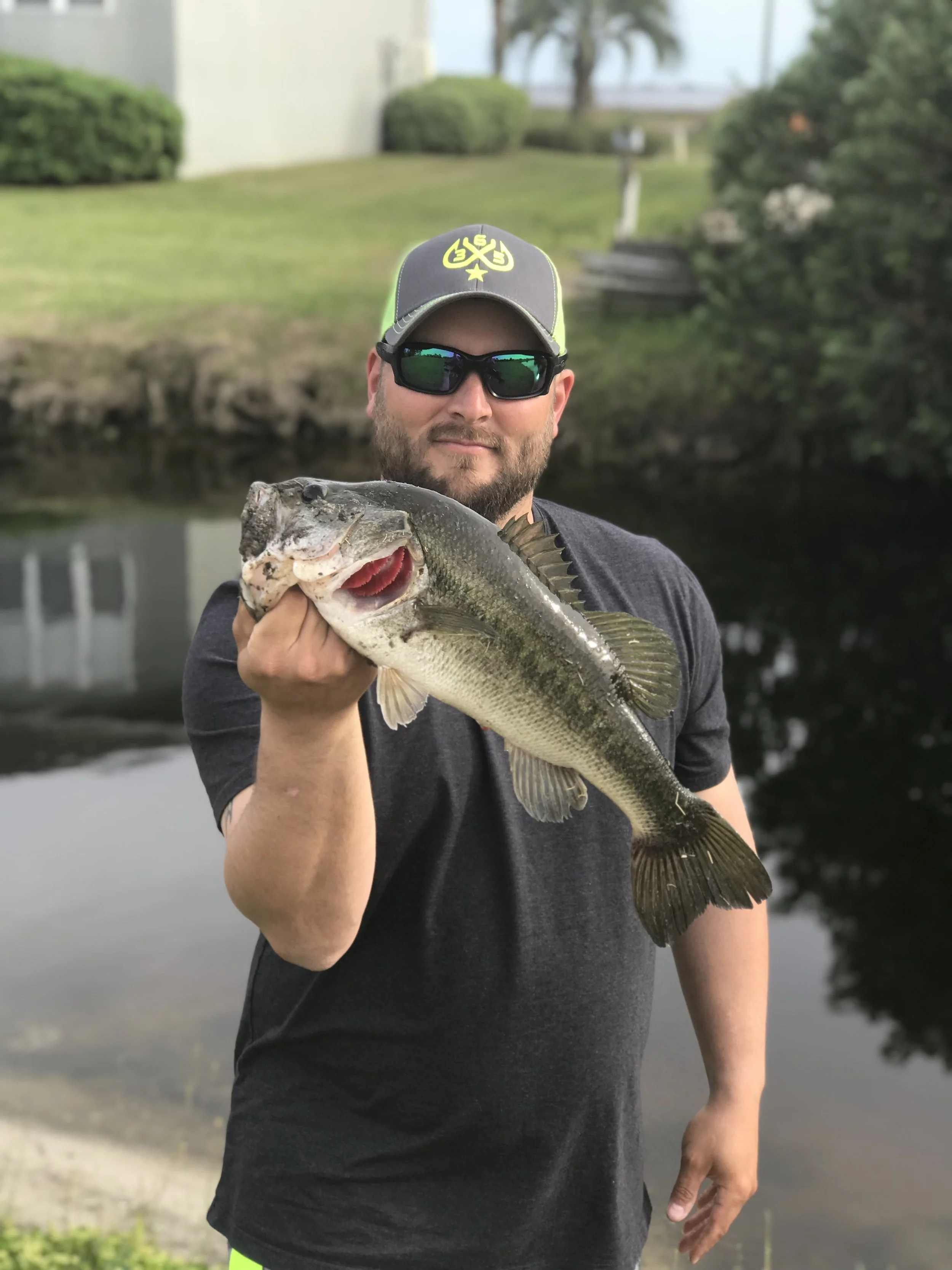 Man holding large fish near a canal with greenery and a house in the background.