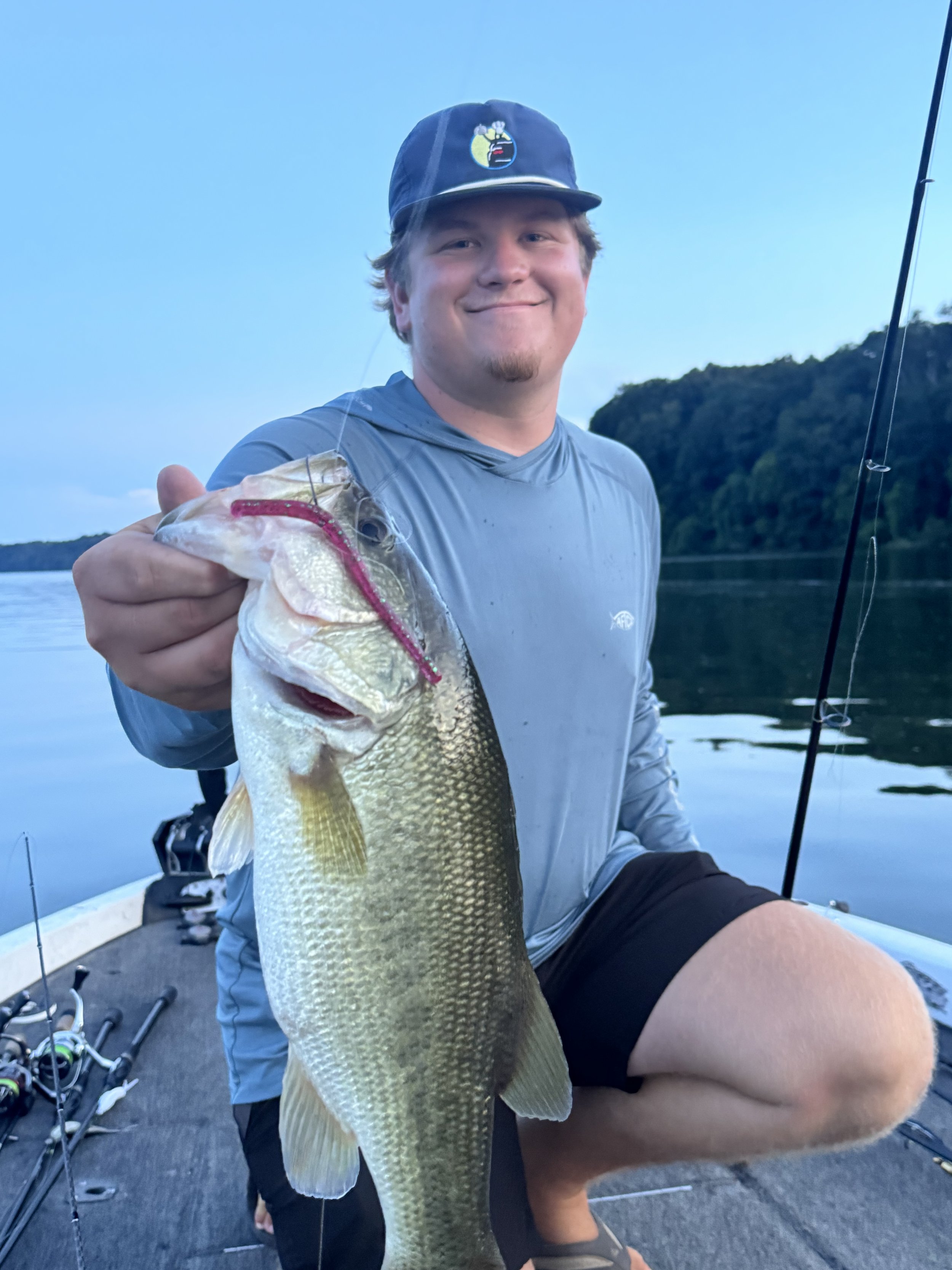 A young man smiling and holding a large fish he caught while sitting on a fishing boat. The background shows a calm body of water and trees.