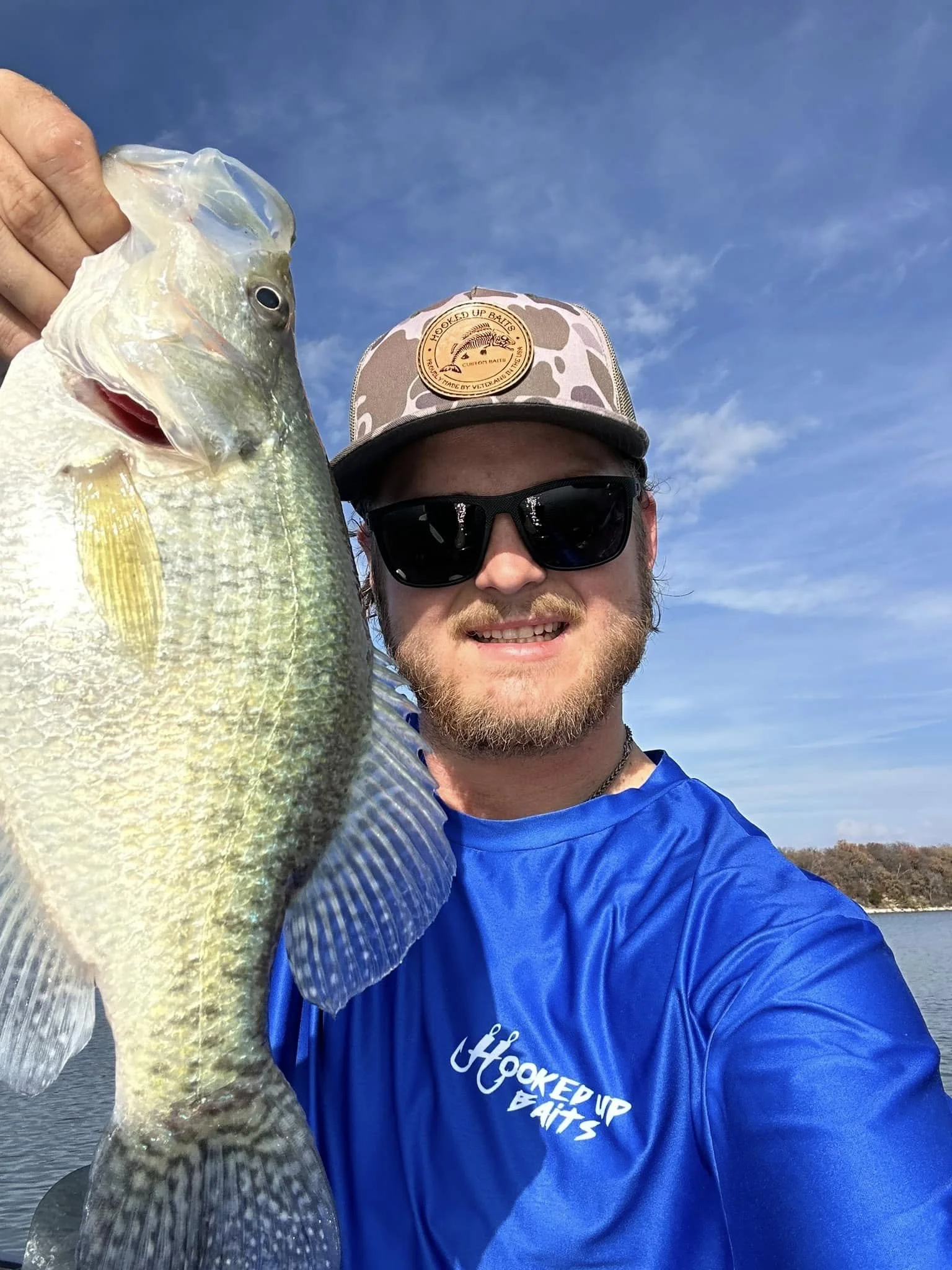 Person holding a large crappie fish, wearing a blue Hooked Up Baits fishing shirt and hat, standing by the water under a clear blue sky.