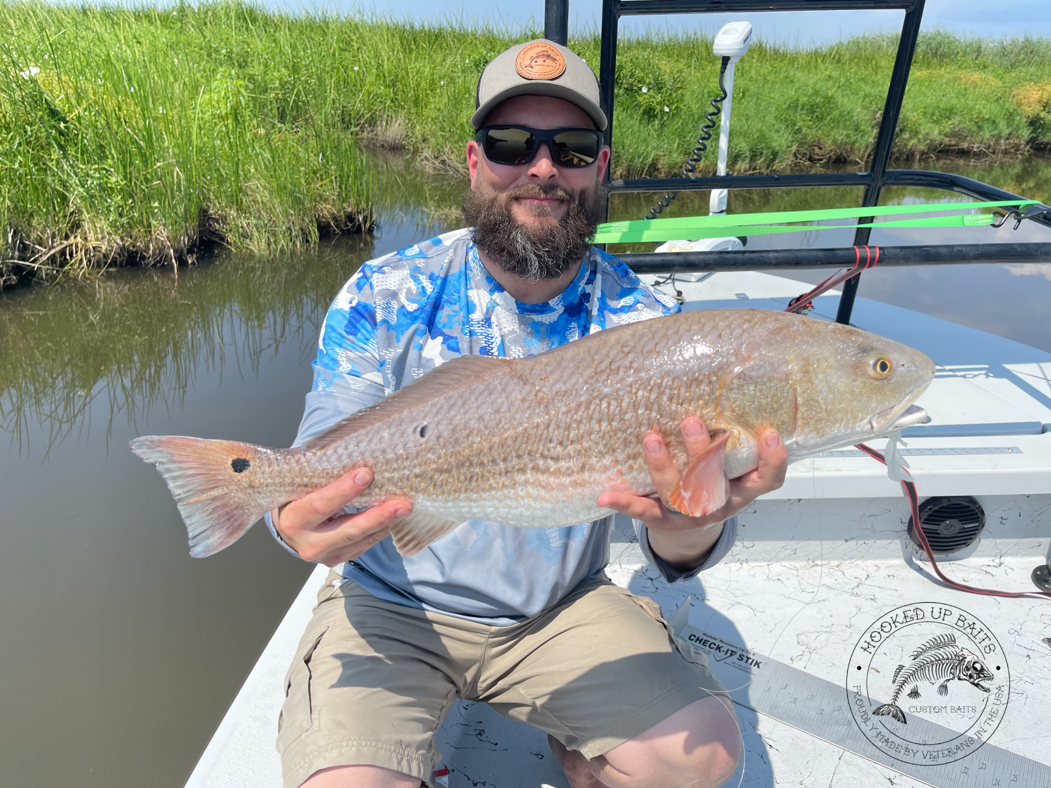 Man holding a large red drum fish on a boat in a marshland setting, wearing sunglasses, a cap, and a blue patterned long-sleeve shirt.