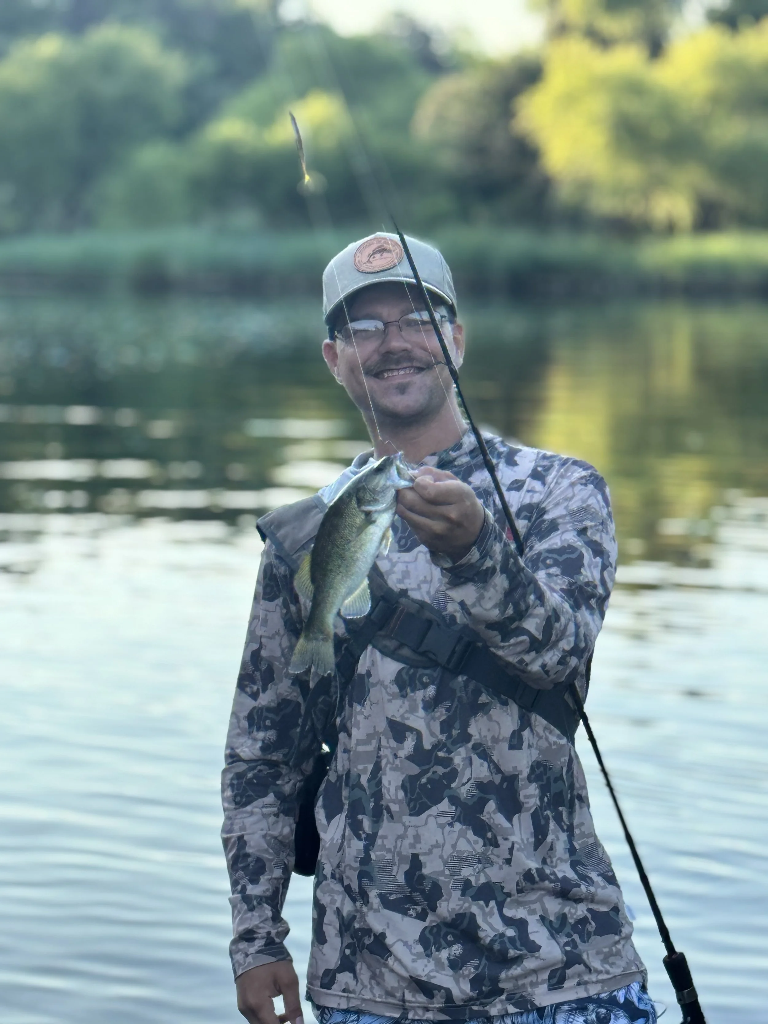 Man in camouflage clothes holding a fish with a fishing rod near a lake during daytime.