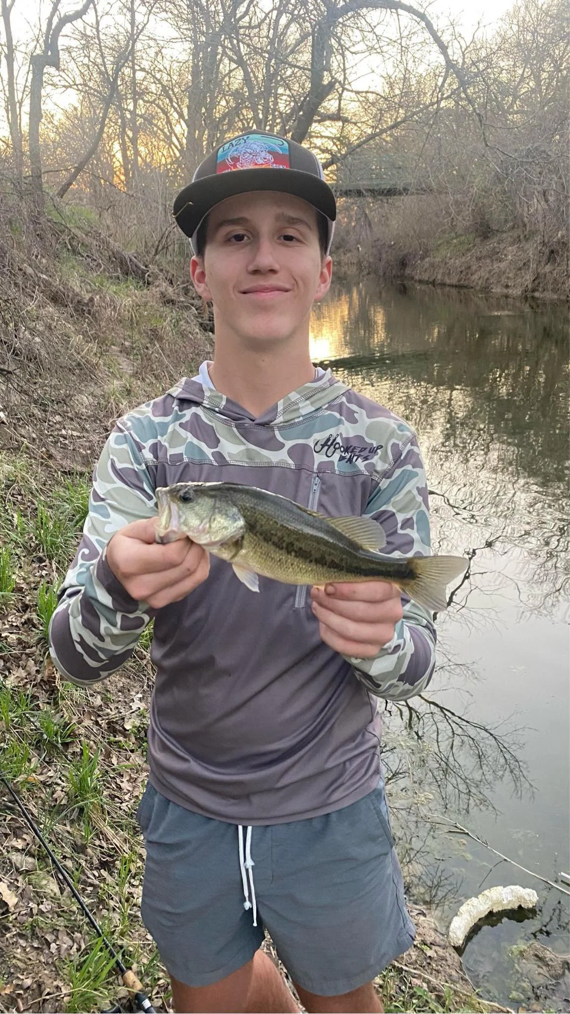 Teenage boy wearing a camouflage hoodie and a cap holds a largemouth bass fish by a river at sunset, with leafless trees and a bridge in the background.