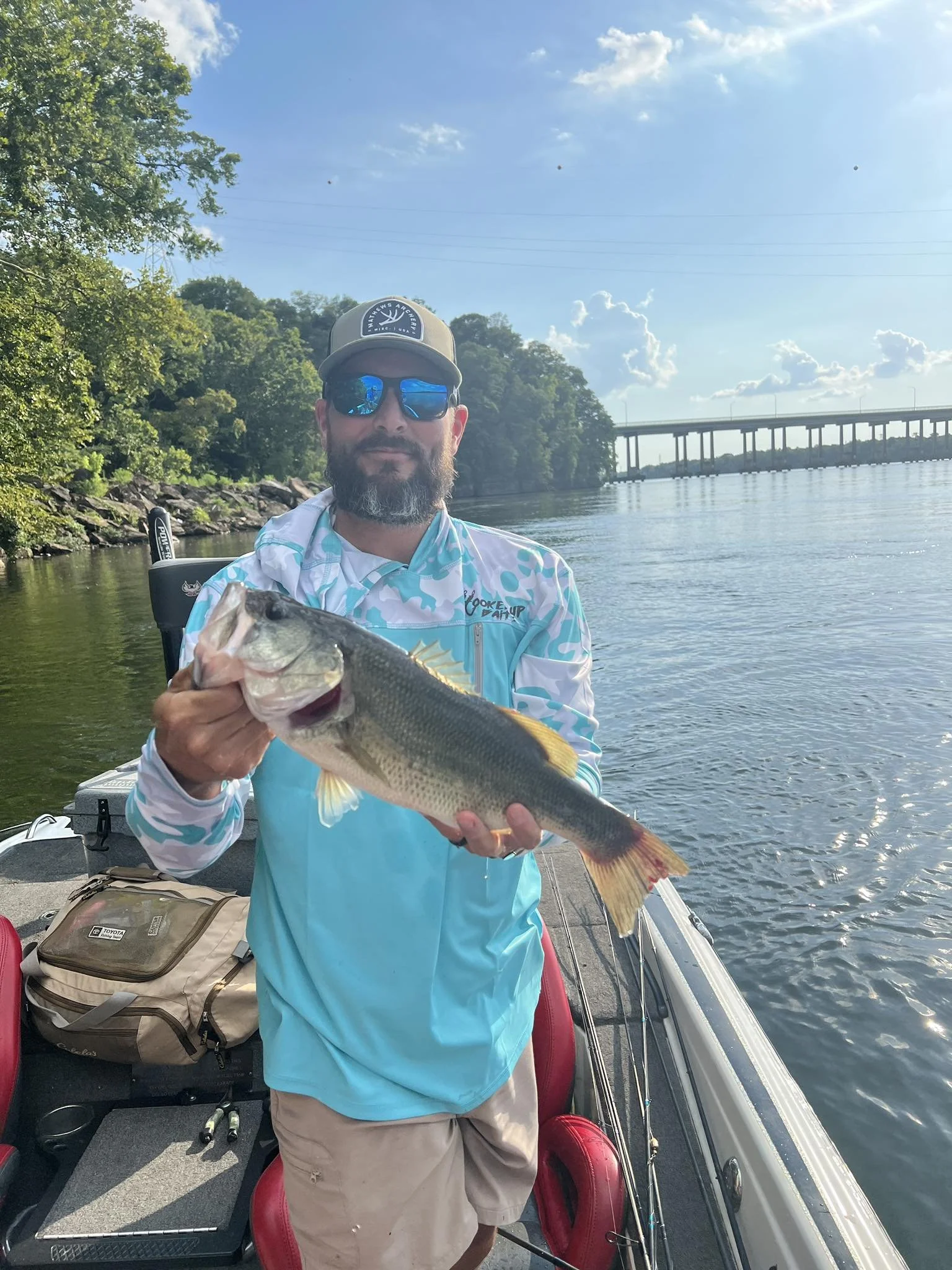Man in sunglasses and a cap holding a largemouth bass fish on a boat.
