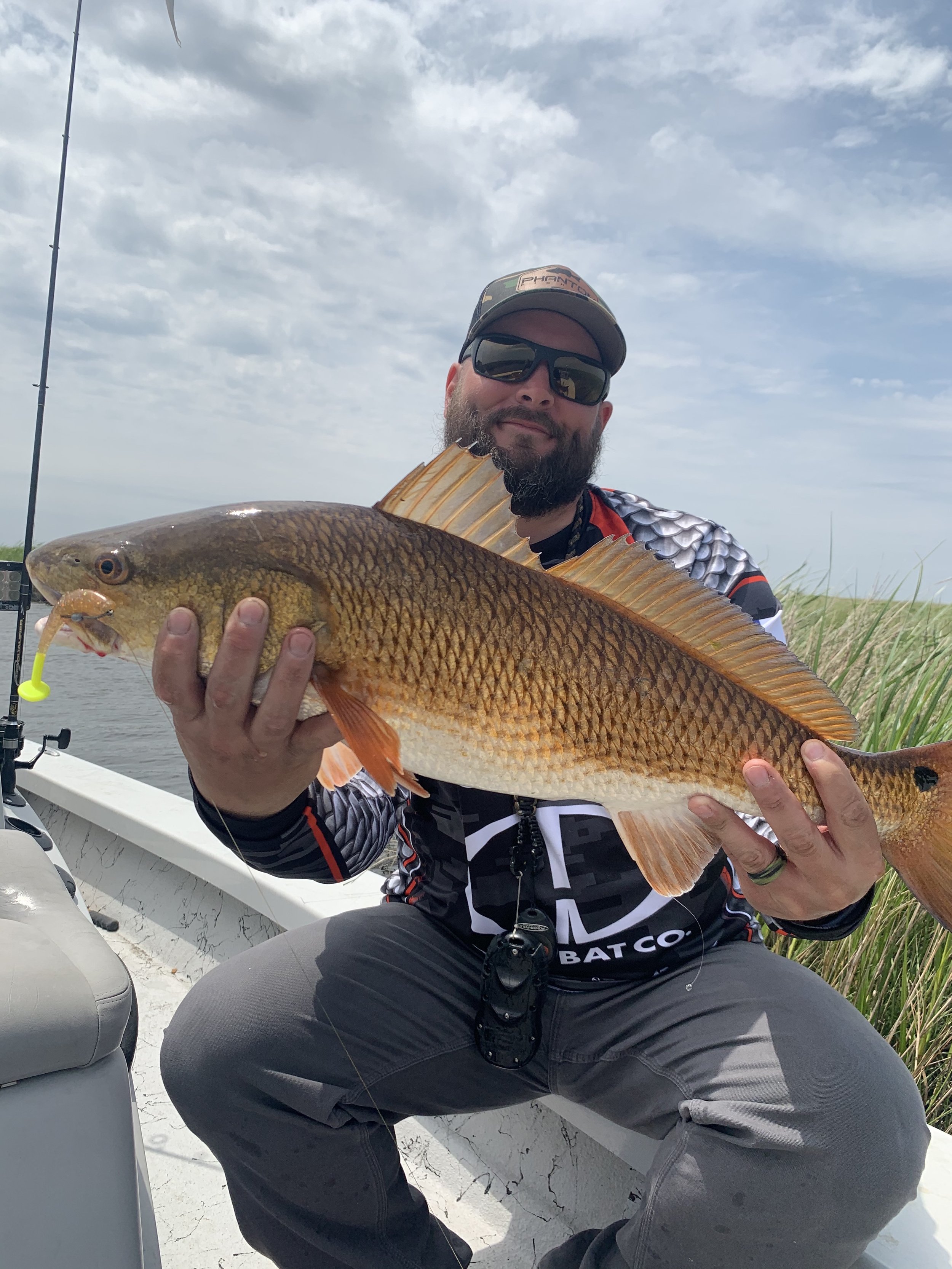 Man holding a large redfish on a boat, wearing sunglasses and a cap, with grassy wetlands in the background.