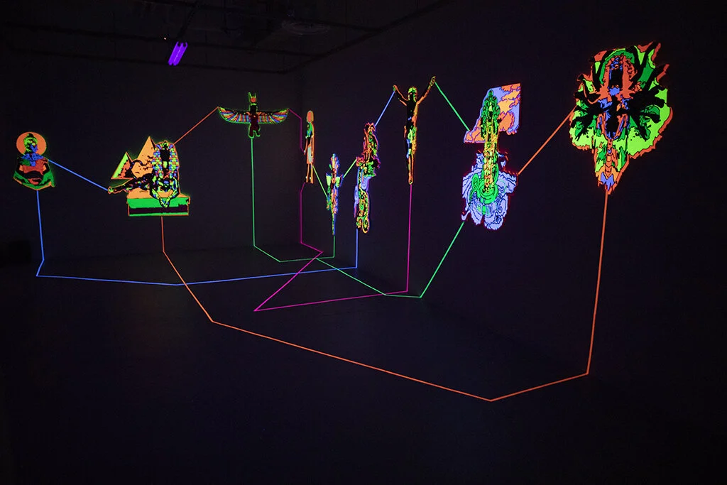 "Emanations" is an installation by artist Nafis Watson depicting deities that have been carved out using a CNC machine. The deities are painted in ultraviolet paint and connected to one another using ultraviolet tape. 