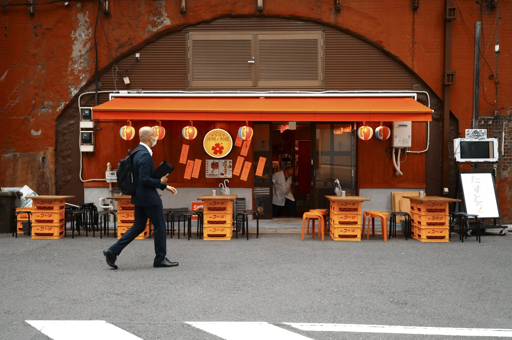 A person in a suit and face mask walking past a small restaurant with orange decorations, including paper lanterns and crates, on an urban street.