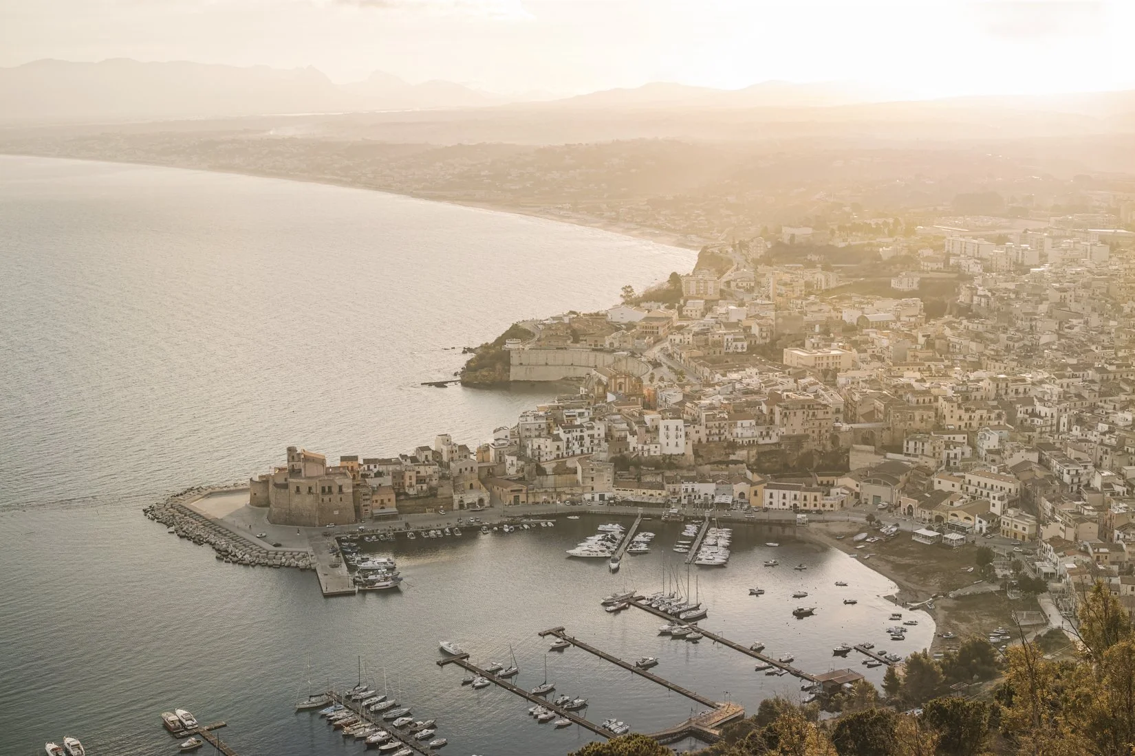 A view of a coastal town with a marina, harbor, and numerous buildings at sunset or sunrise.