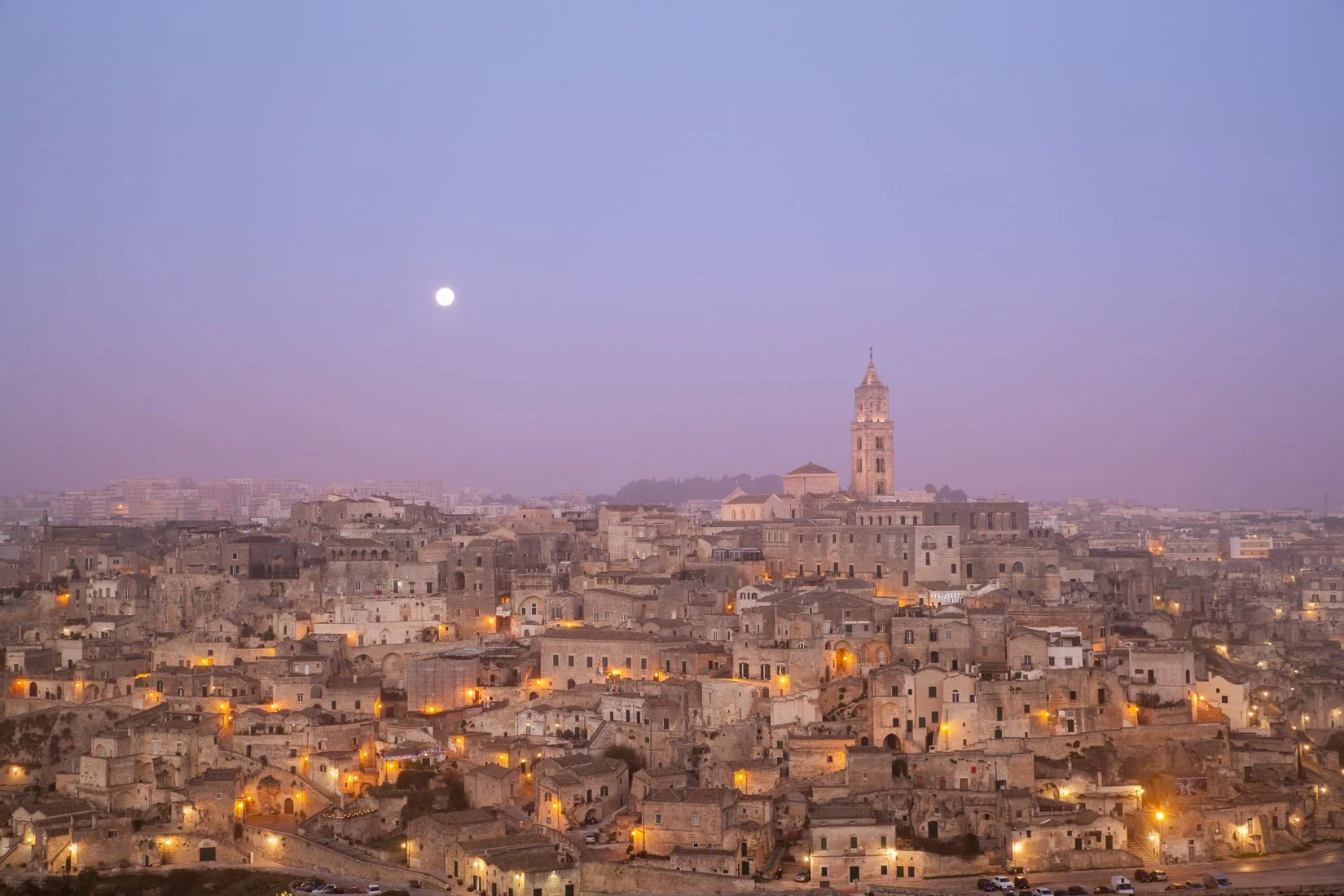 Cityscape of an ancient Italian town at dusk, with numerous stone buildings, a prominent tower, and a visible moon in the sky.