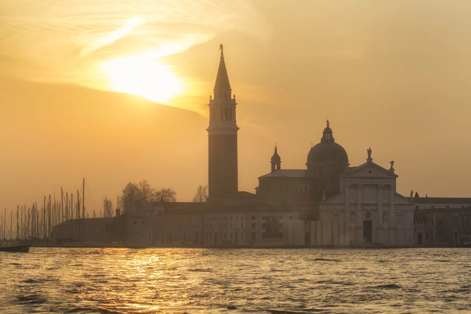 Sunset over Venice with silhouette of a church and boat masts in the water.