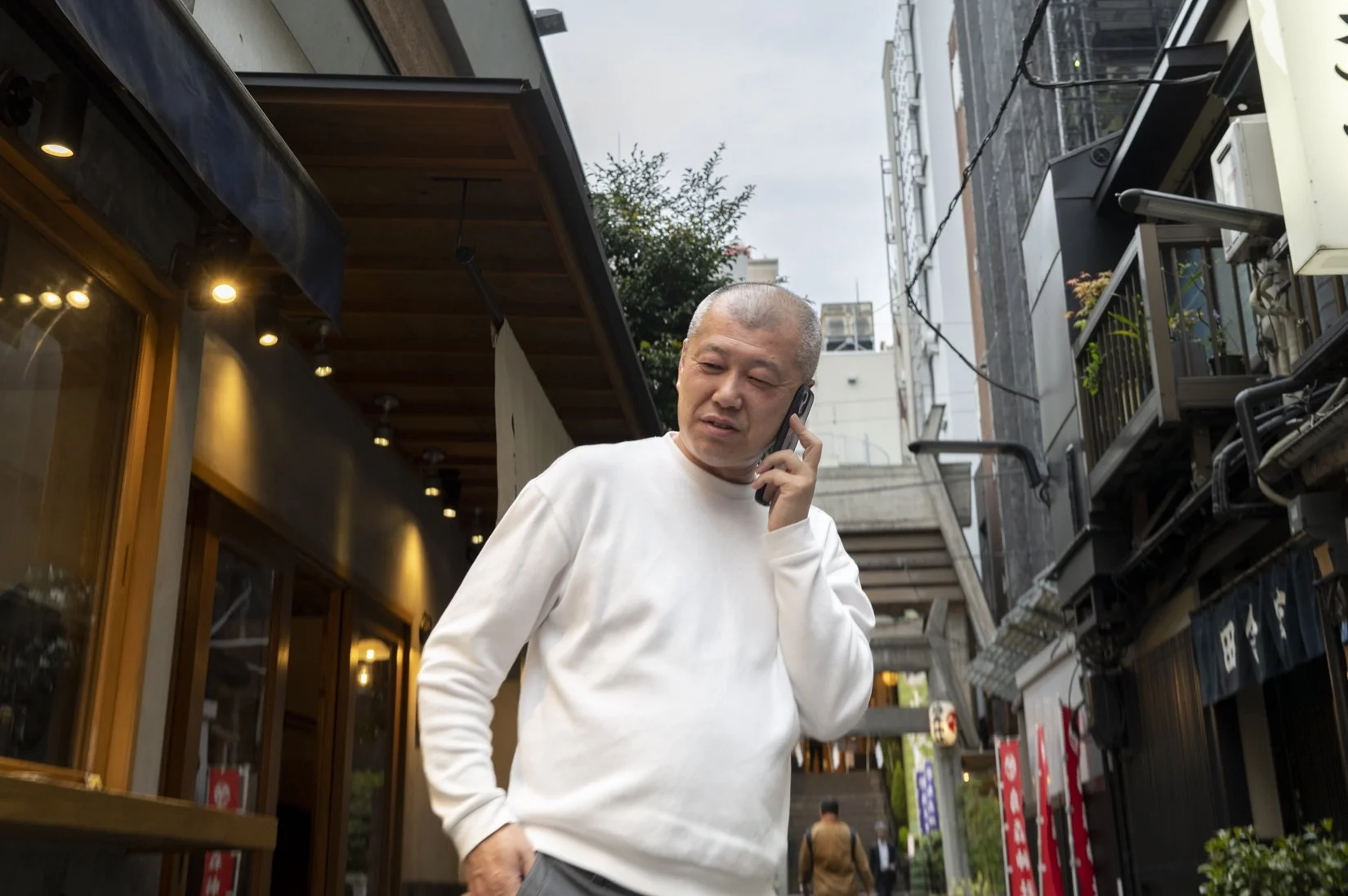 An older man with short gray hair, wearing a white long-sleeve shirt, is talking on a cellphone on a city street. He looks slightly confused or concerned.