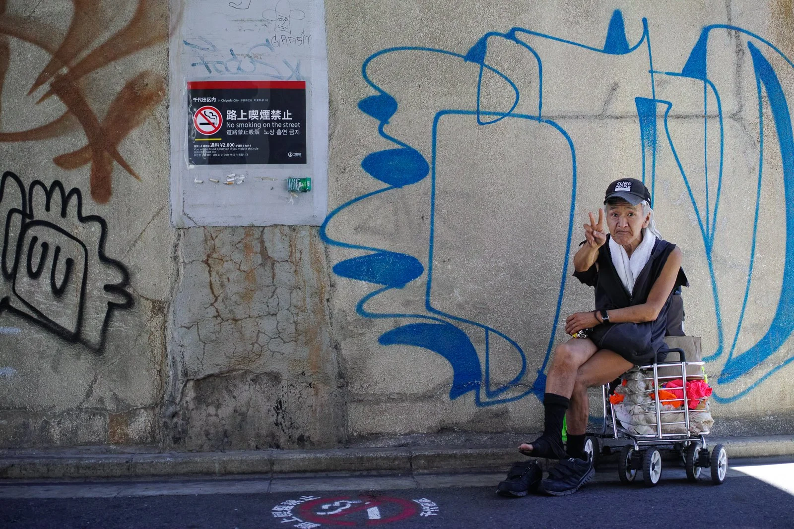 An elderly person with a bandage on the head sitting in a small cart on a sidewalk, making a peace sign with their hand. The background includes graffiti on a wall and a 'no smoking' sign.