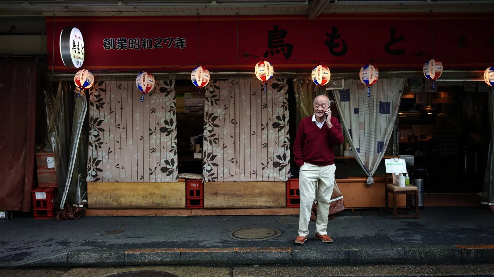 An elderly man stands outside a Japanese restaurant, talking on his cellphone. The restaurant has red hanging lanterns, a red sign with Japanese characters, and curtains with a leaf pattern at the entrance.