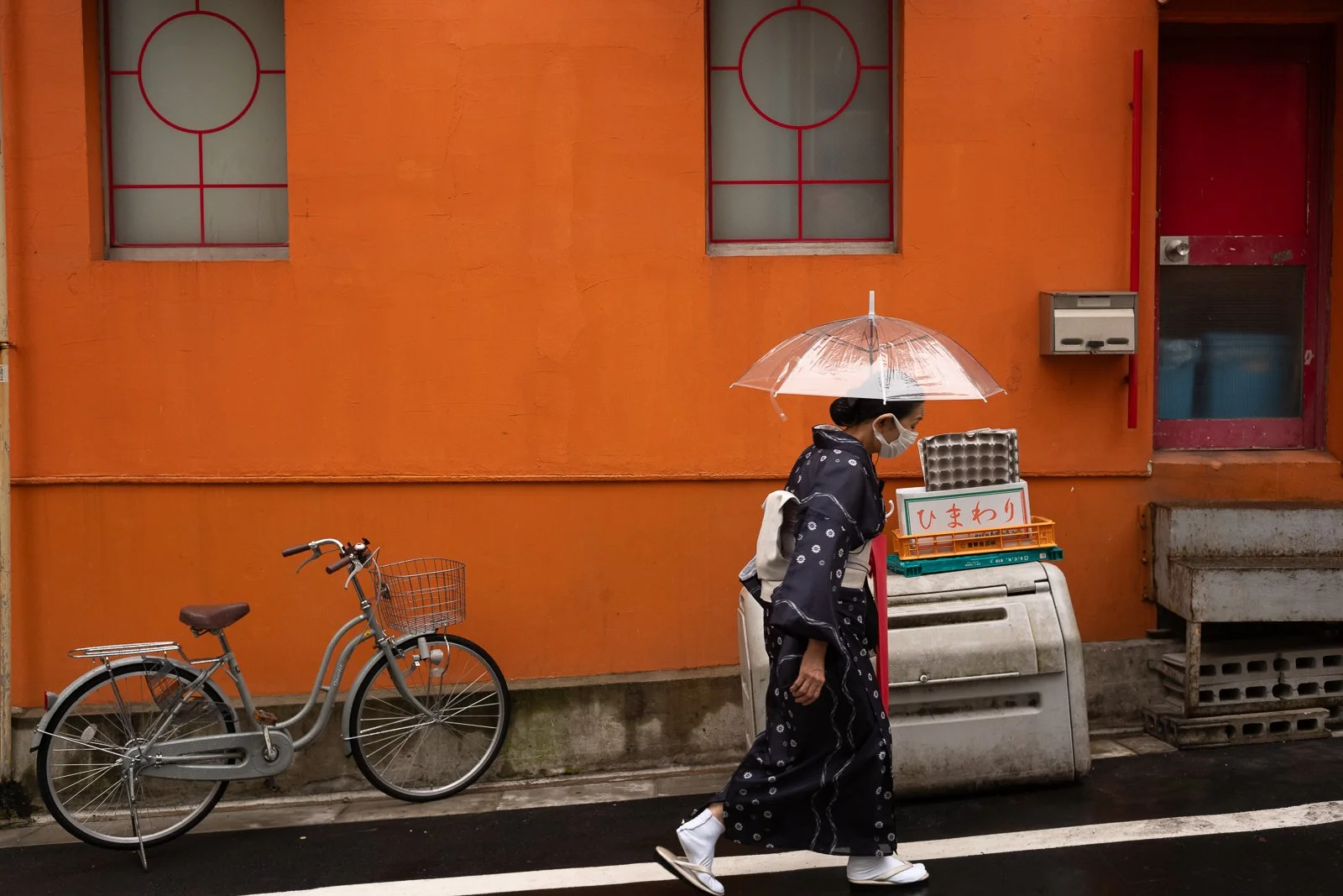 A woman dressed in traditional Japanese kimono walking on a wet street holding a transparent umbrella, with a bicycle parked against an orange wall and a box with Japanese writing.