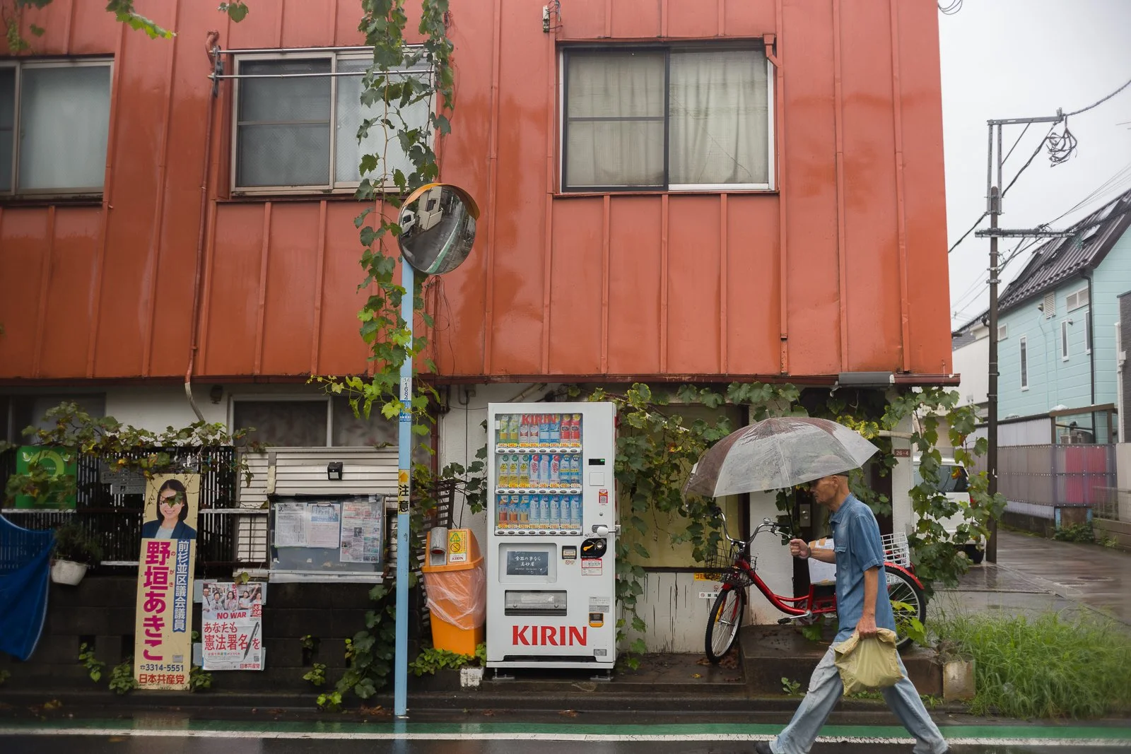 Pedestrian carrying a transparent umbrella past a red building and vending machine on a rainy Tokyo street.
