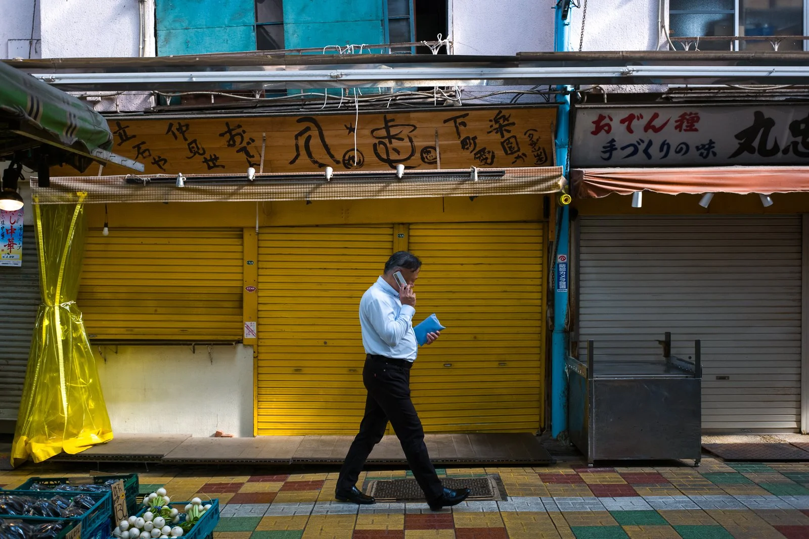 A man in business attire walking down a colorful sidewalk while talking on his cellphone, holding a notebook, with closed shop fronts in the background.