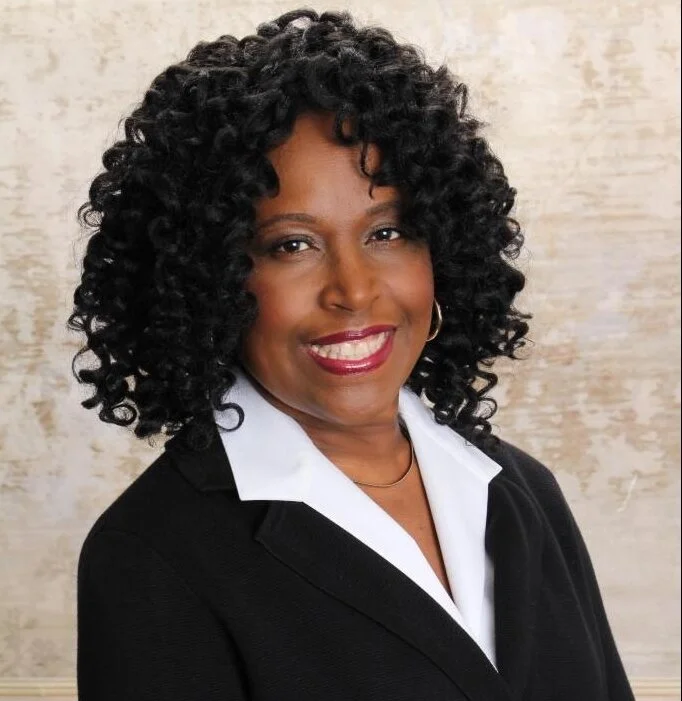 Portrait of a woman with curly black hair, wearing a black blazer and white shirt, smiling, standing against a beige textured background.