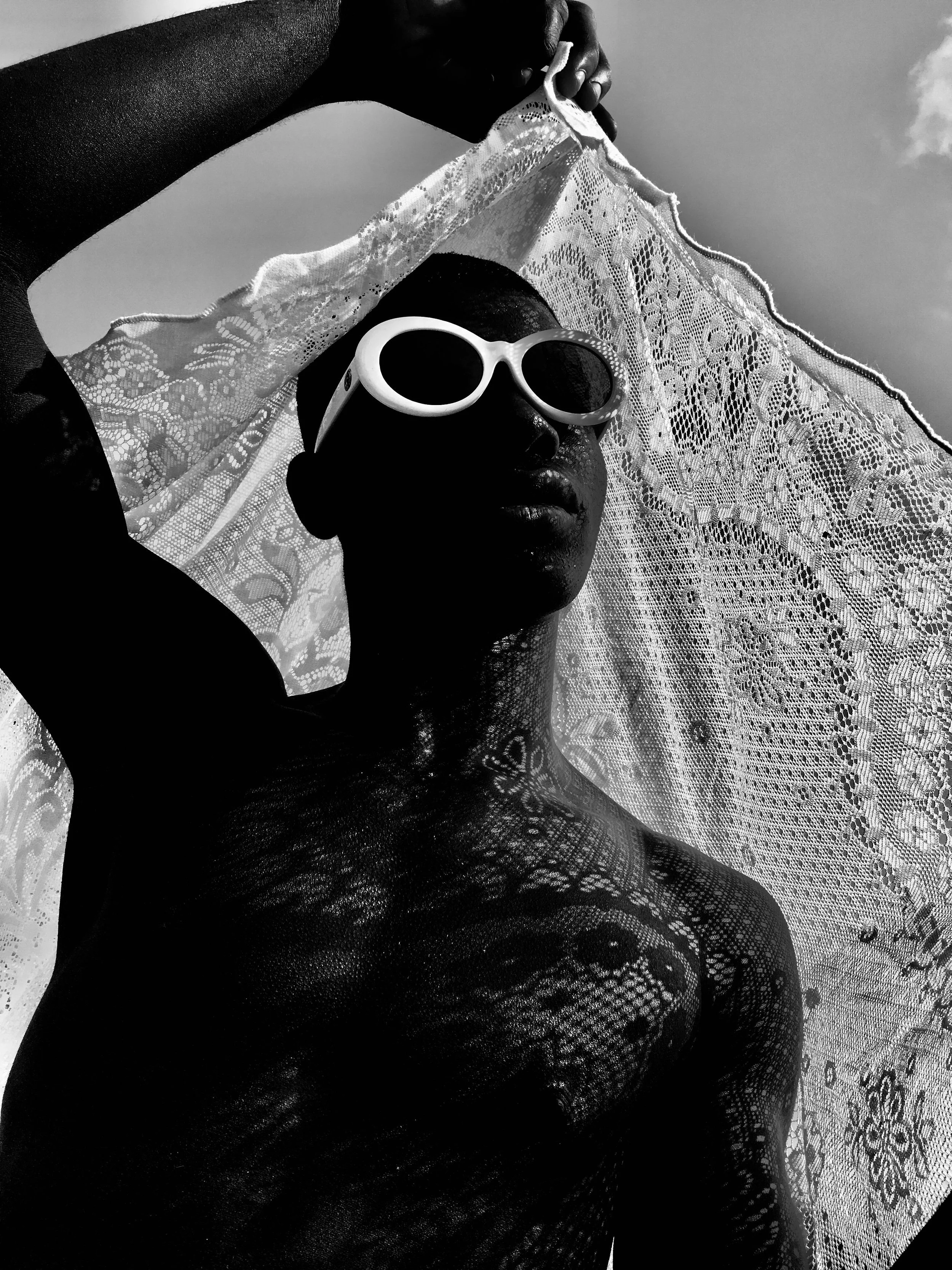 Black and white photo of a person wearing large sunglasses, holding a lace fabric above their head, with intricate shadows cast on their face and body, against a cloudy sky background.