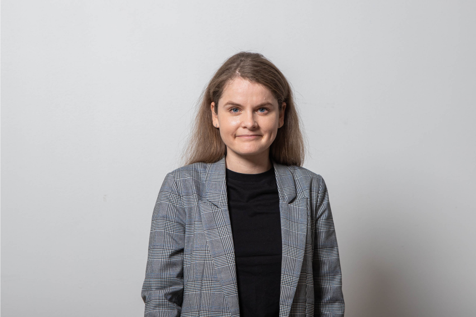 A young woman with light brown hair and blue eyes stands against a plain light gray background, wearing a gray plaid blazer over a black shirt, smiling slightly.
