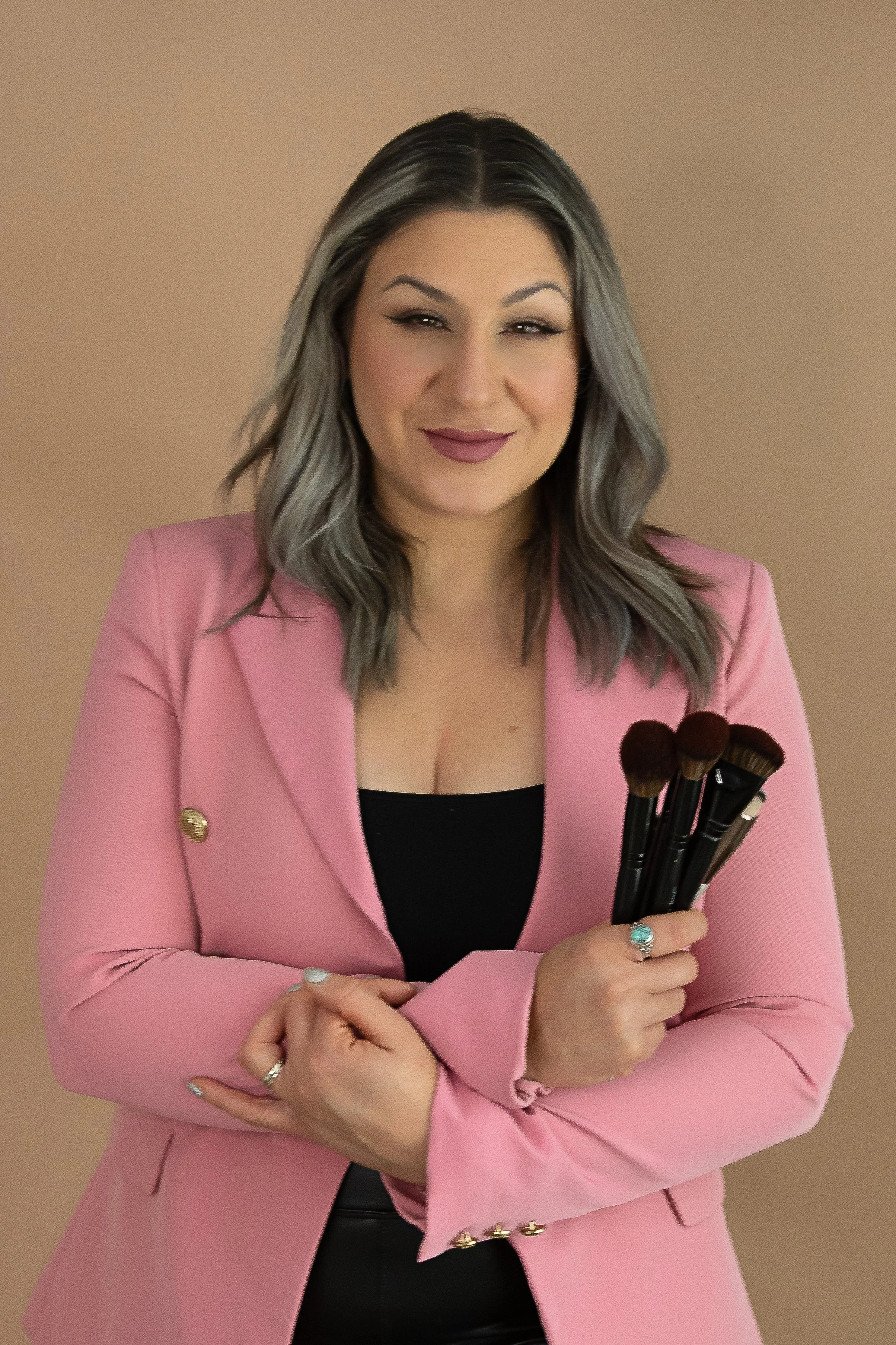 A woman with grayish hair in a pink blazer holding makeup brushes, smiling at the camera against a beige background.