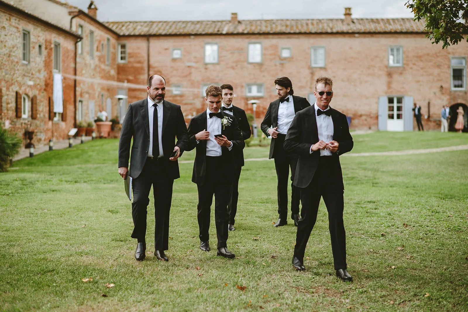 Group of men in custom made Aleksandar Stately tuxedos walking on grass outside a brick building, smiling, at a formal event.