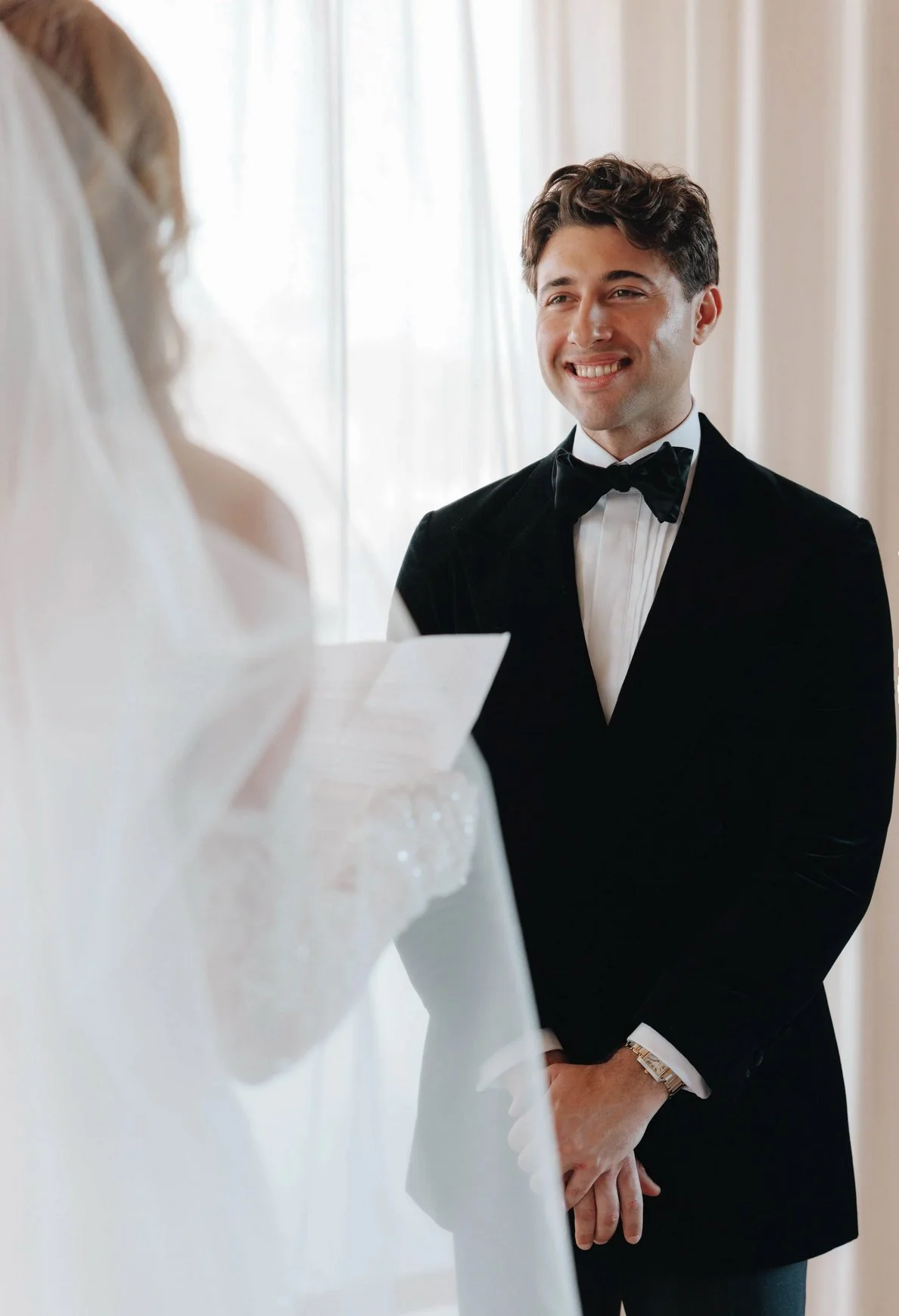 A groom in a black Aleksandar Stately custom made to measure tuxedo and bow tie stands smiling during a wedding ceremony.