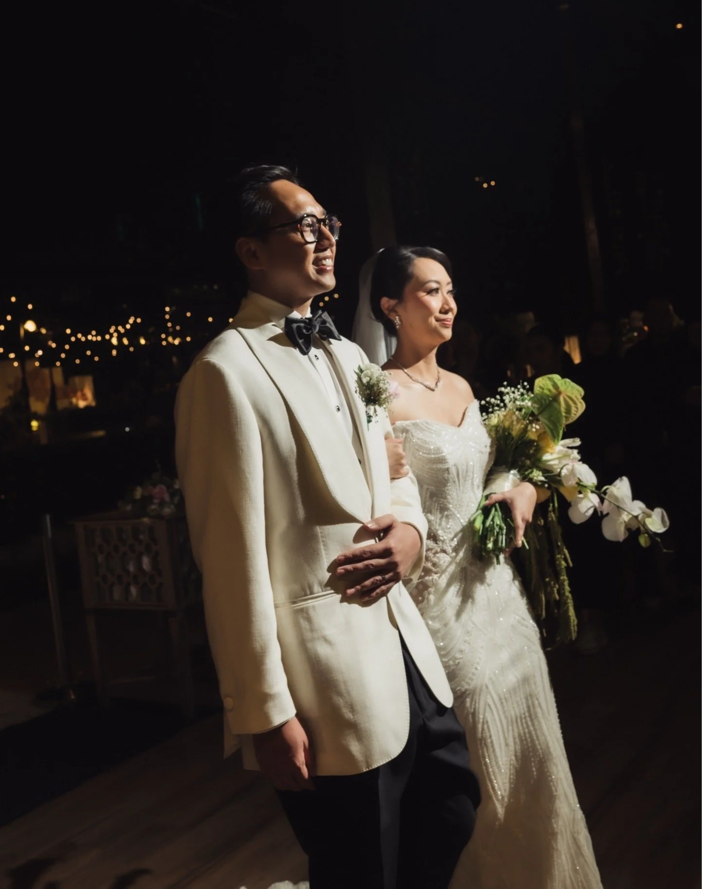 A bride and groom dressed in custom tailor made Aleksandar Stately Suit, standing side by side during a wedding reception, both dressed in formal attire, with the bride holding a bouquet of flowers.