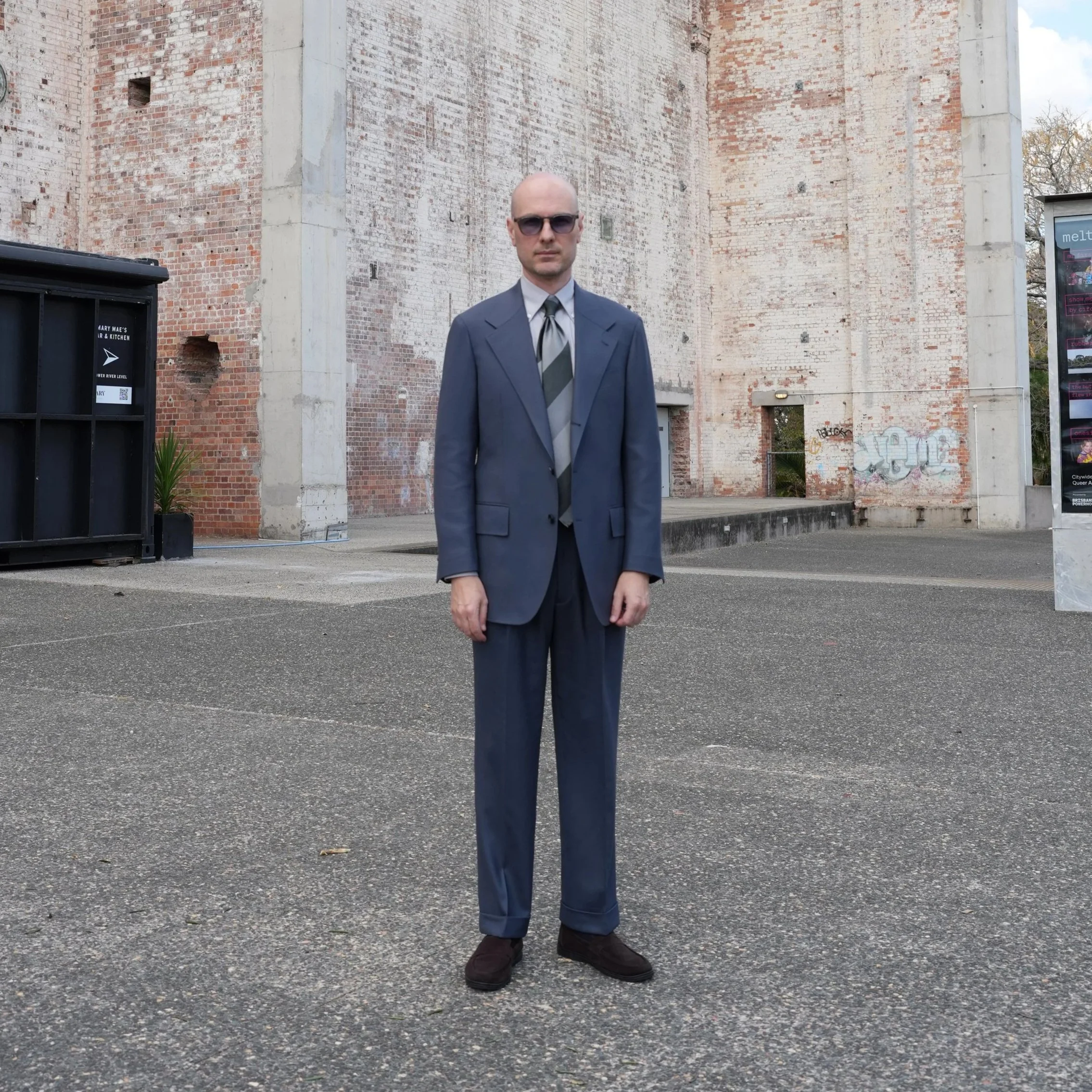 Aleksandar Stately wearing a custom tailor made to measure blue suit, and a striped tie stands outdoors in Brisbane.
