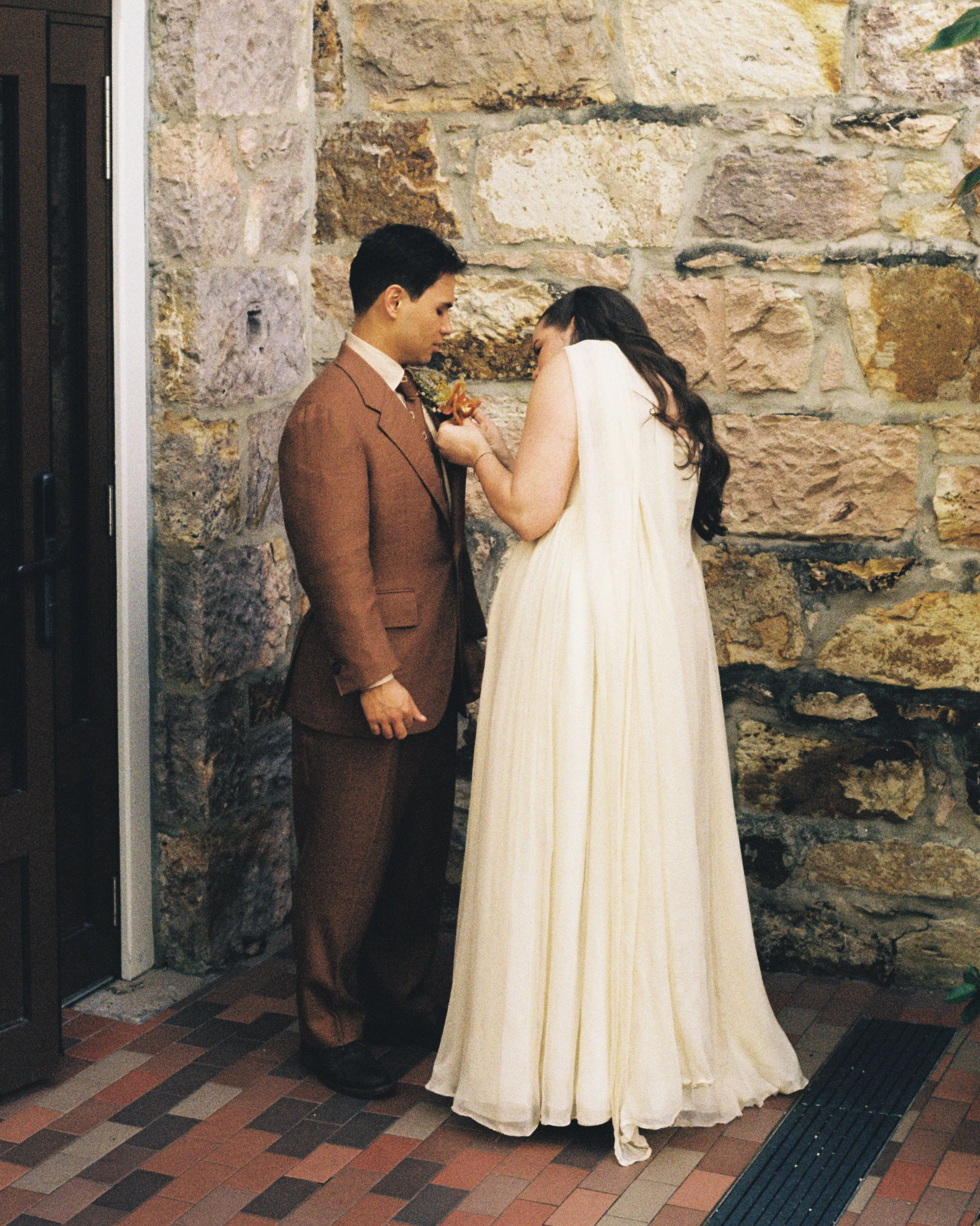A man in a brown Aleksandar Stately Made to measure wedding suit and a woman in a white gown are standing close together, facing each other against a stone wall. They appear to be sharing an intimate moment; the woman is touching the man's chest.