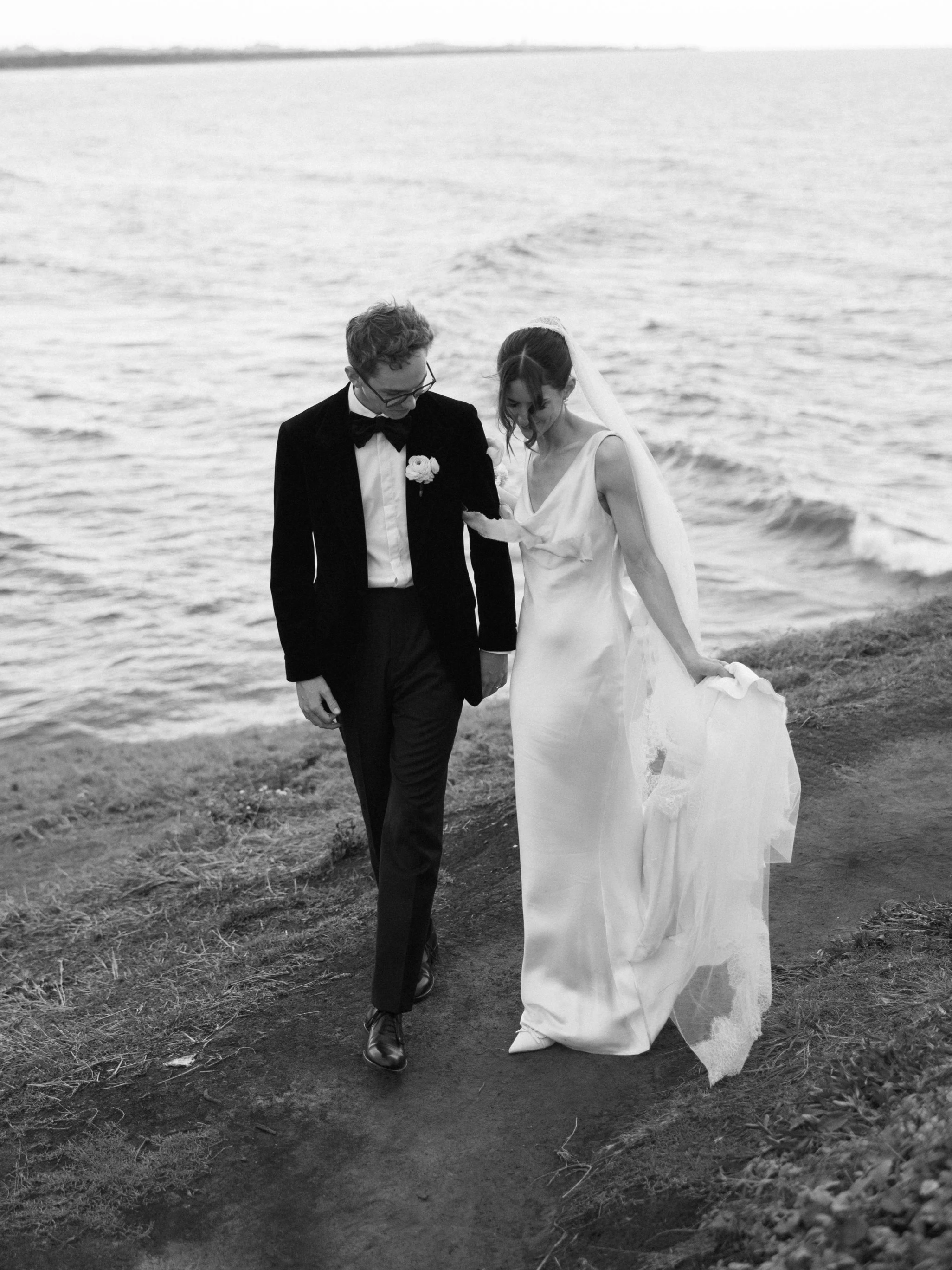 A Aleksandar Stately made to measure Suit black-and-white photo of a bride and groom walking along a shoreline holding hands, with the bride in a white wedding dress and the groom in a tuxedo.
