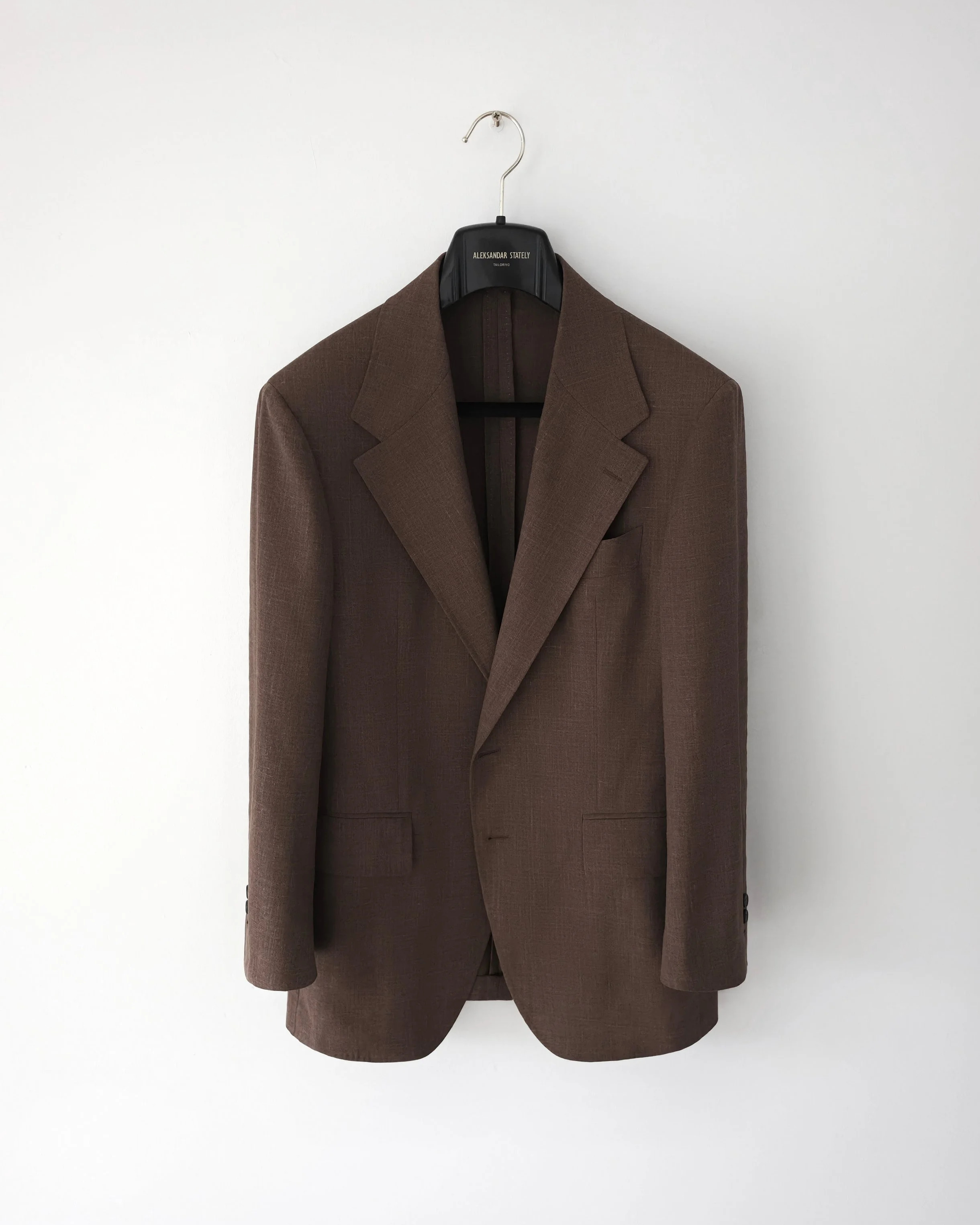 Brown tailored blazer hanging on a black hanger with the label 'ALEKSANDAR STATELY' against a plain white wall.