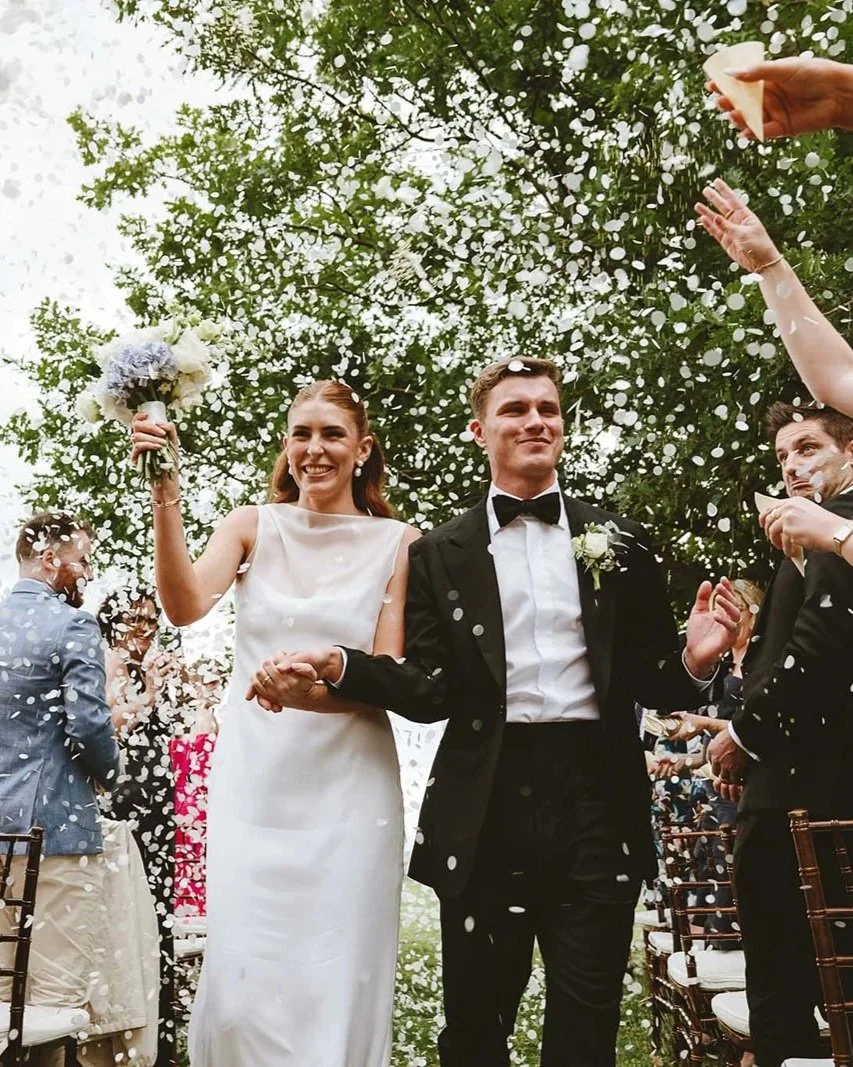 A bride and groom walking down the aisle outdoors, celebrating their wedding with guests throwing confetti. The bride is holding a bouquet and smiling, while the groom is dressed in a Aleksandar Stately custom made tuxedo with a white boutonniere.