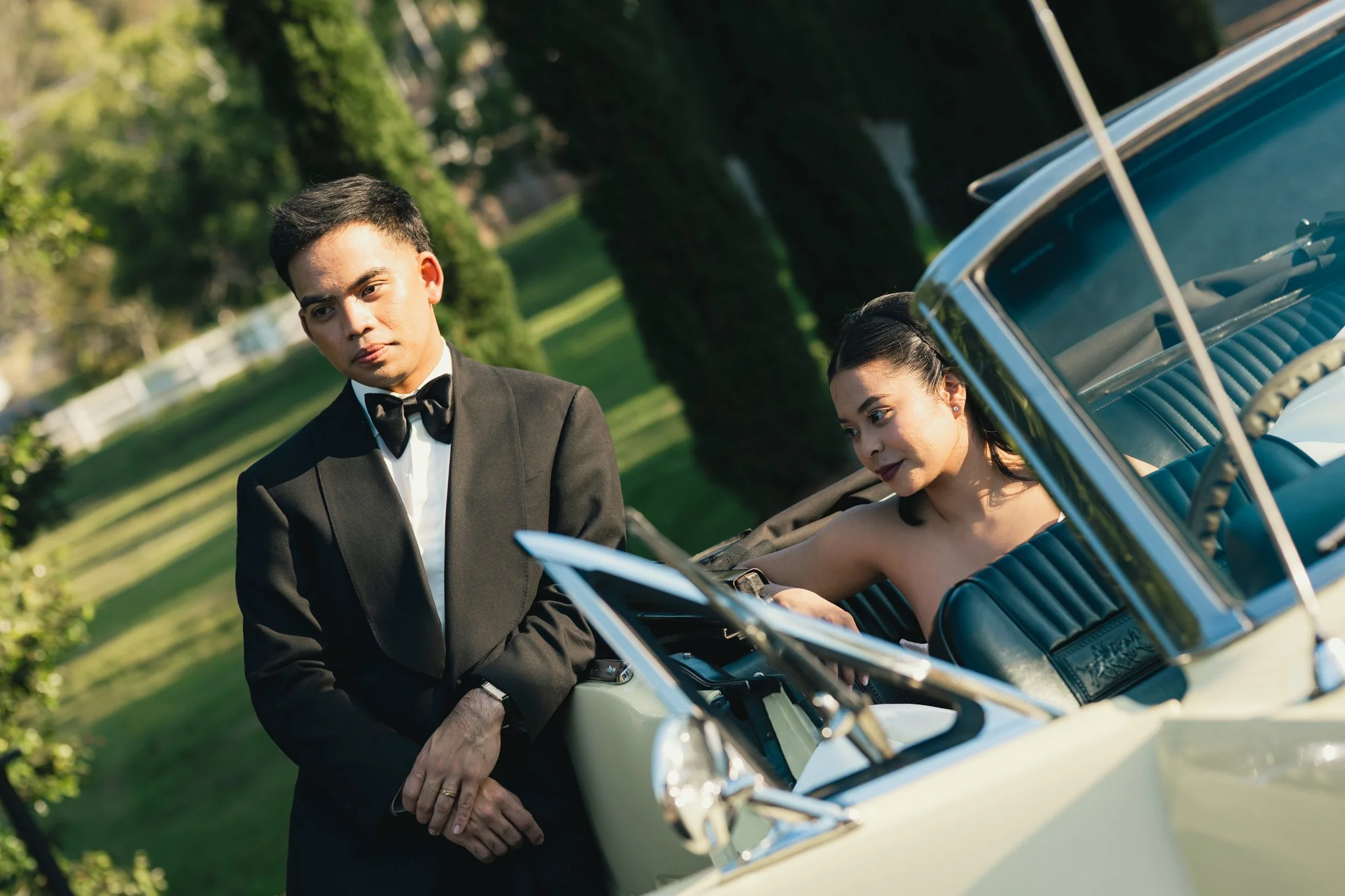 A man in a made to measure custom tailored Aleksandar Stately tuxedo standing next to a woman in a dress sitting in a vintage car in a park-like setting.
