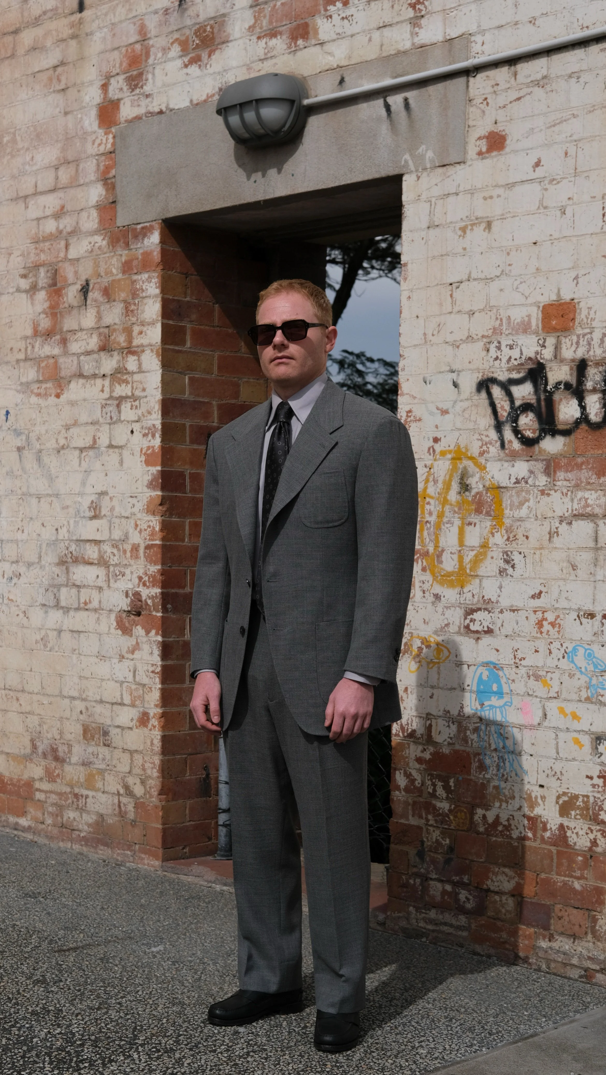 A man wearing a grey Aleksandar Stately custom tailored made to measure suit, hand made Aleksandar Stately grey patterned tie, and sunglasses standing in front of a graffiti-covered brick wall in Brisbane Australia.