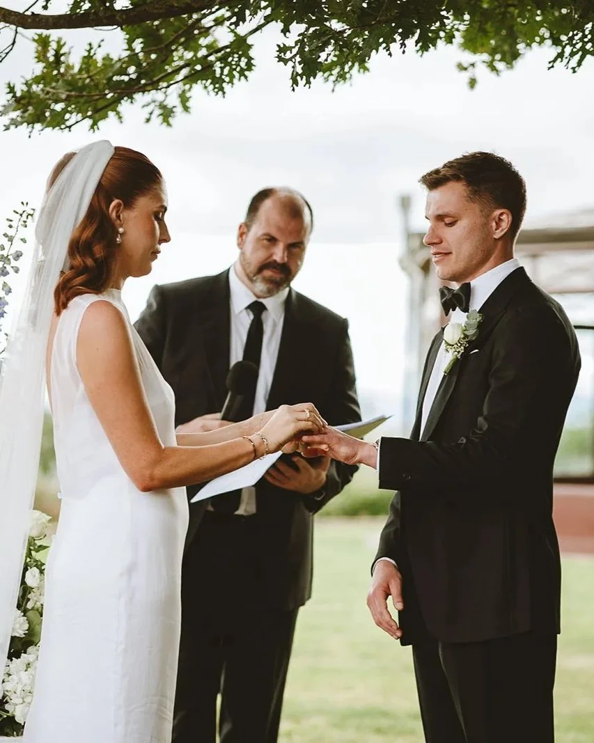 A groom wearing custom made Aleksandar Stately suit exchanging wedding vows outdoors, with an officiant in the background holding a paper, during a wedding ceremony.