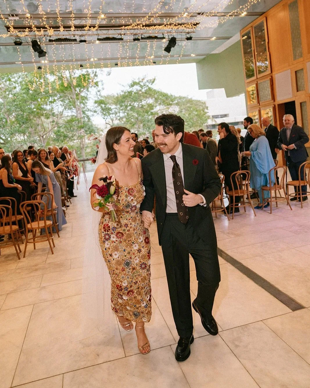 People celebrating a wedding at QPAC Brisbane, with a bride in a floral gown holding a bouquet and a groom wearing a Aleksandar Stately custom tailored made to measure Olive Green Suit and Tie.