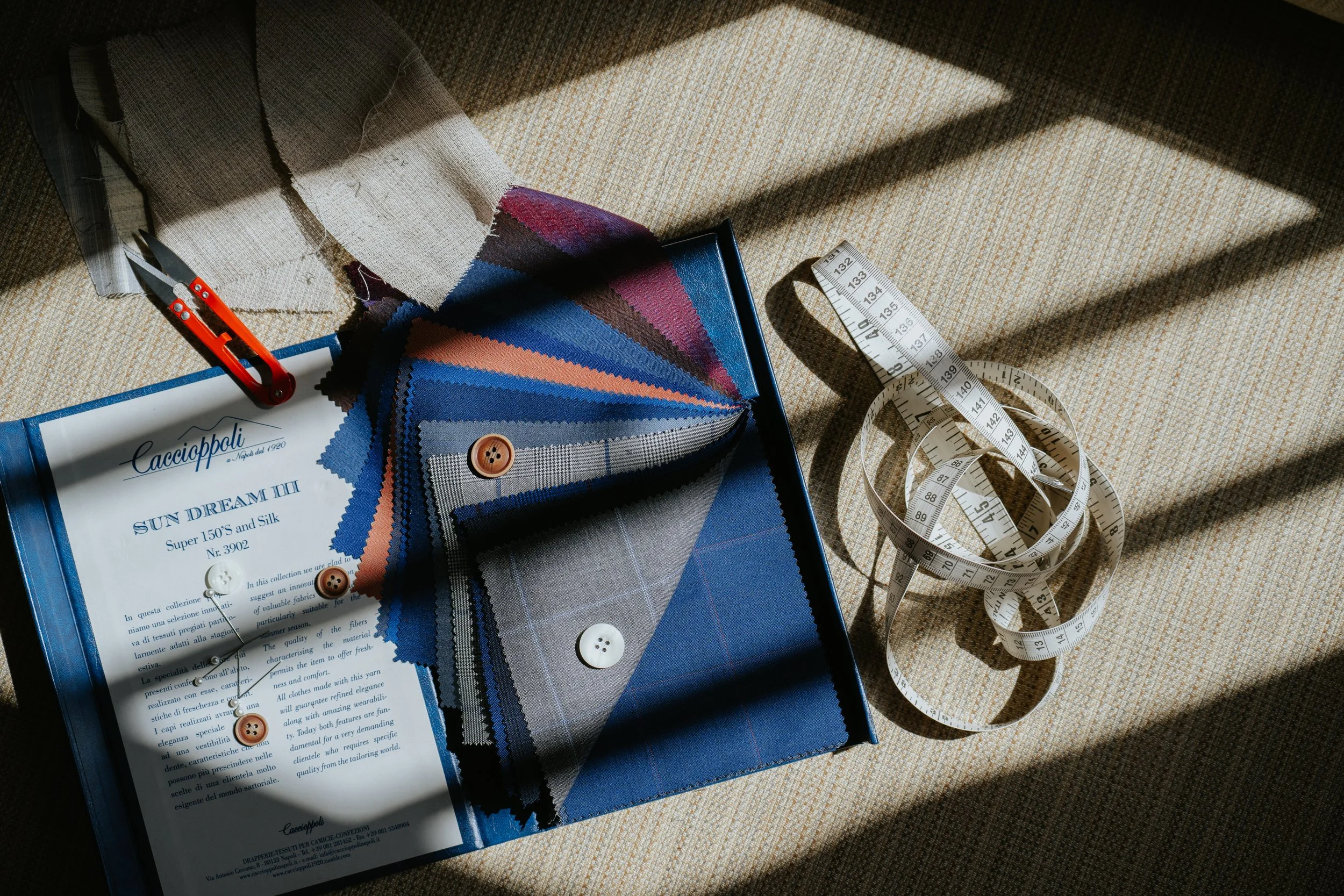 ALEKSANDAR STATELY Fabric swatches, a white measuring tape, a pair of scissors, and fabric buttons placed on a beige carpet, with sunlight casting shadows across the scene.
