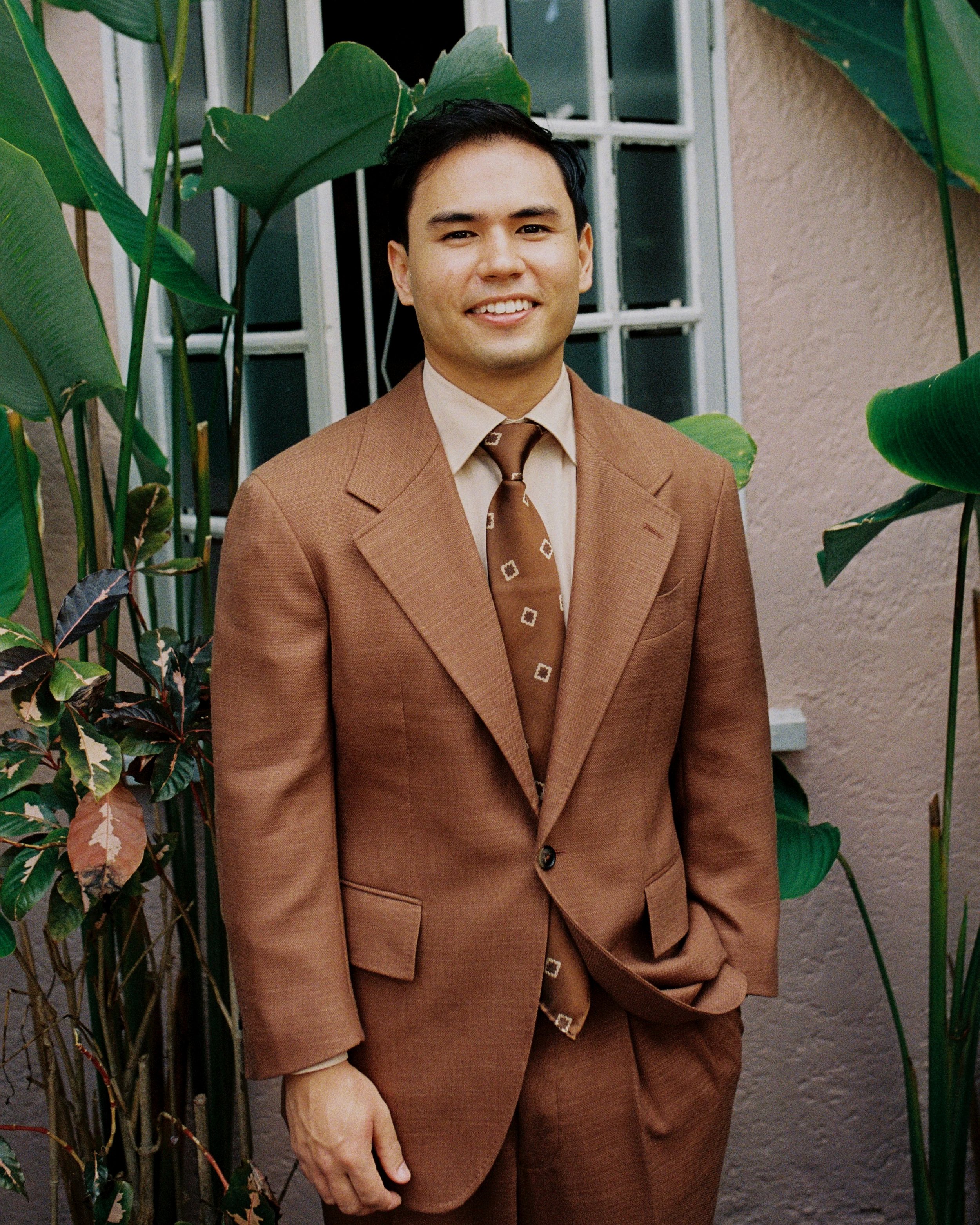 A man in a brown Aleksandar Stately made to measure wedding suit standing outdoors near a wall with large green plants.