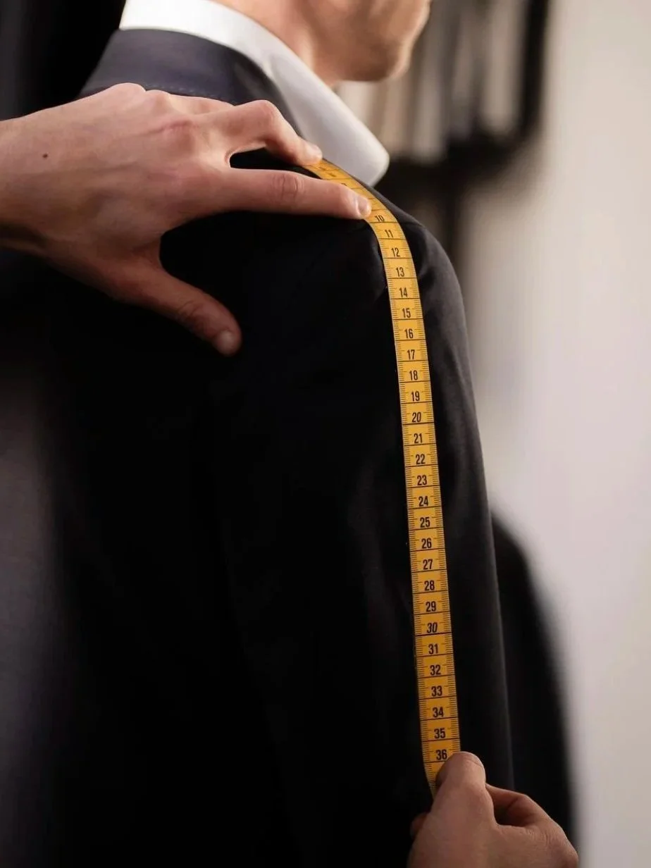 Person's shoulder being measured with a yellow tape measure, wearing a black suit and white shirt.
