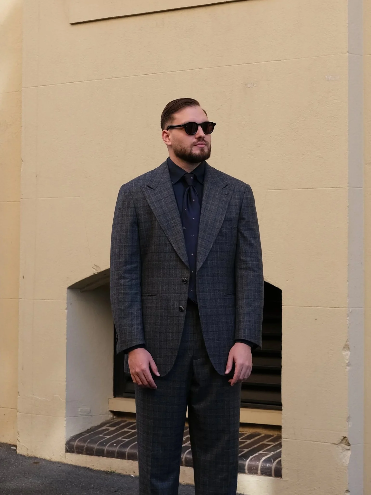 A man with a beard and slicked back hair wearing a custom tailored made to measure Aleksandar Stately dark shirt, a blue tie, a gray plaid suit.