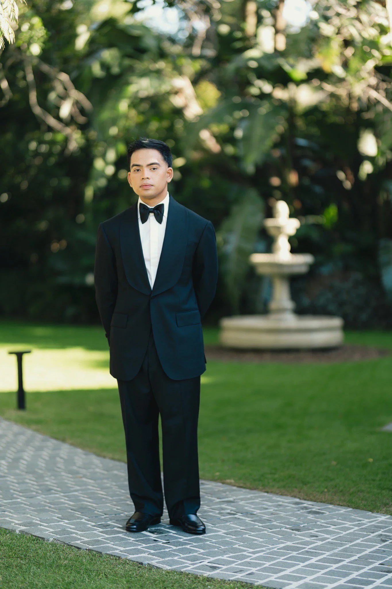 A young man in made to measure custom tailored Aleksandar Stately tuxedo, standing on a brick pathway in a garden with lush greenery and a stone fountain in the background.
