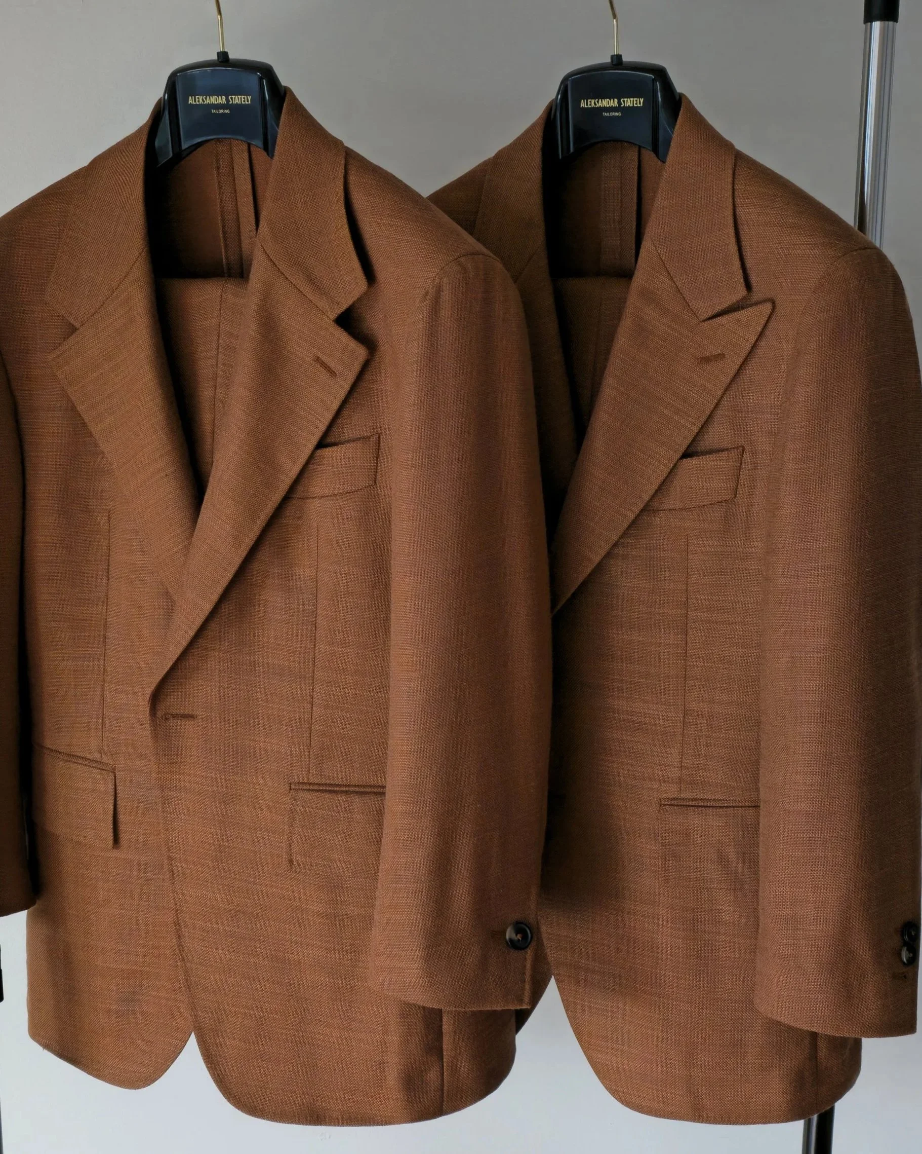 Two Aleksandar Stately made to measure brown tailored blazers hanging side by side on black hangers labeled 'Aleksandar Statly TAILORING'.