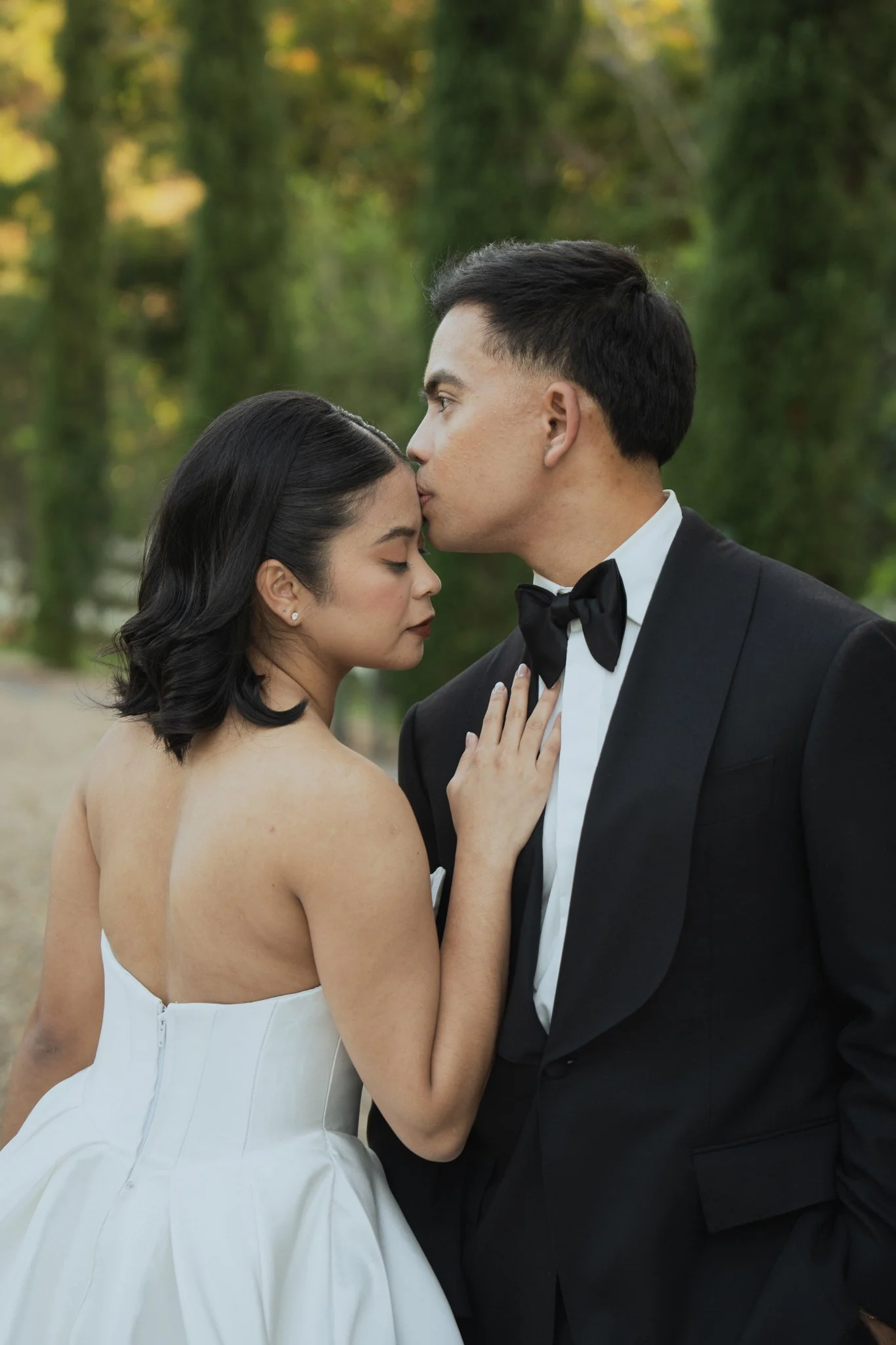 A man in a made to measure custom tailored Aleksandar Stately tuxedo and a woman in a wedding dress standing close, with the man kissing the woman's forehead in an outdoor setting with greenery in the background.
