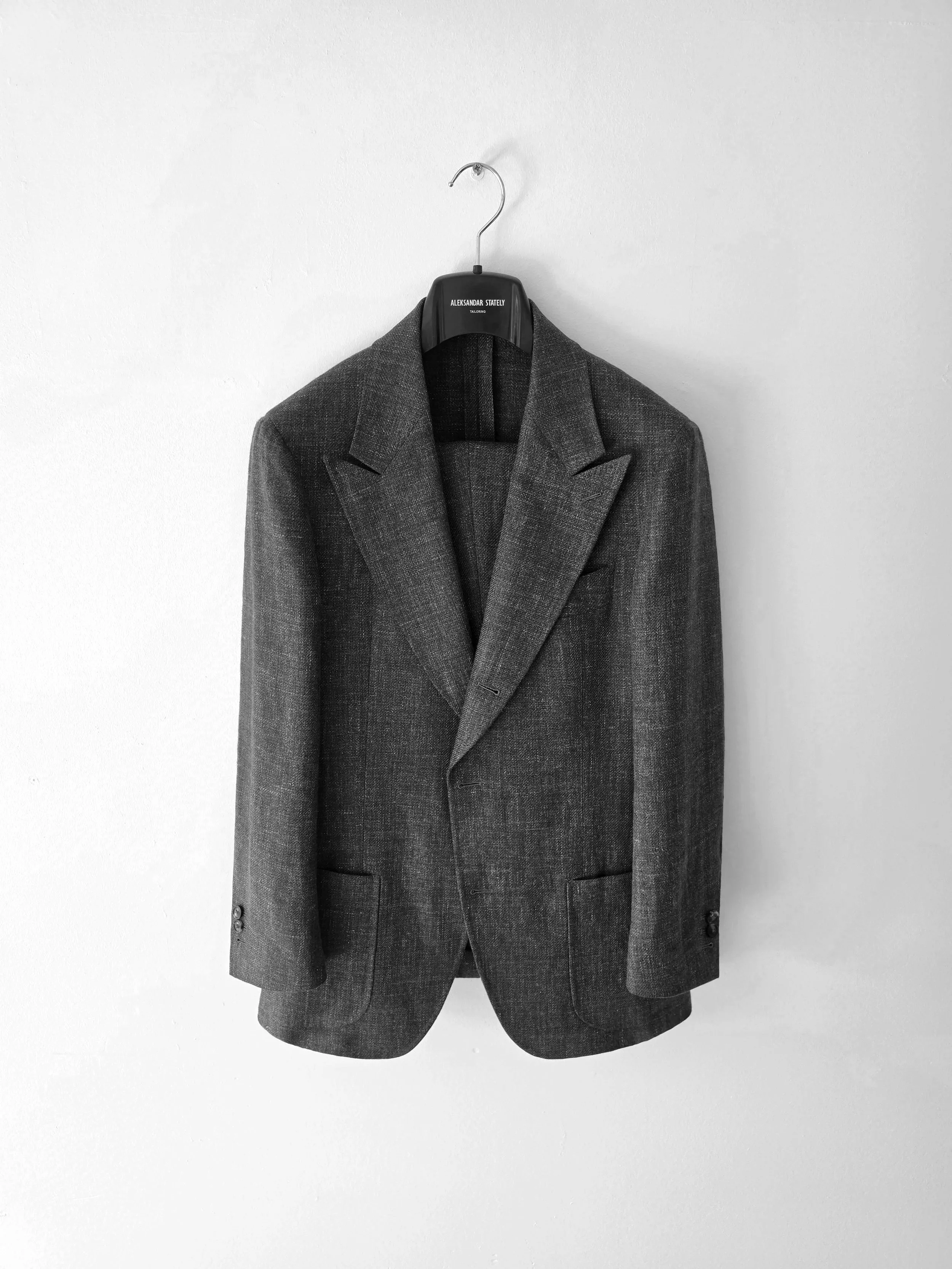 A Aleksandar Stately made to measure suit jacket hanging on a clothing hanger against a plain white wall.