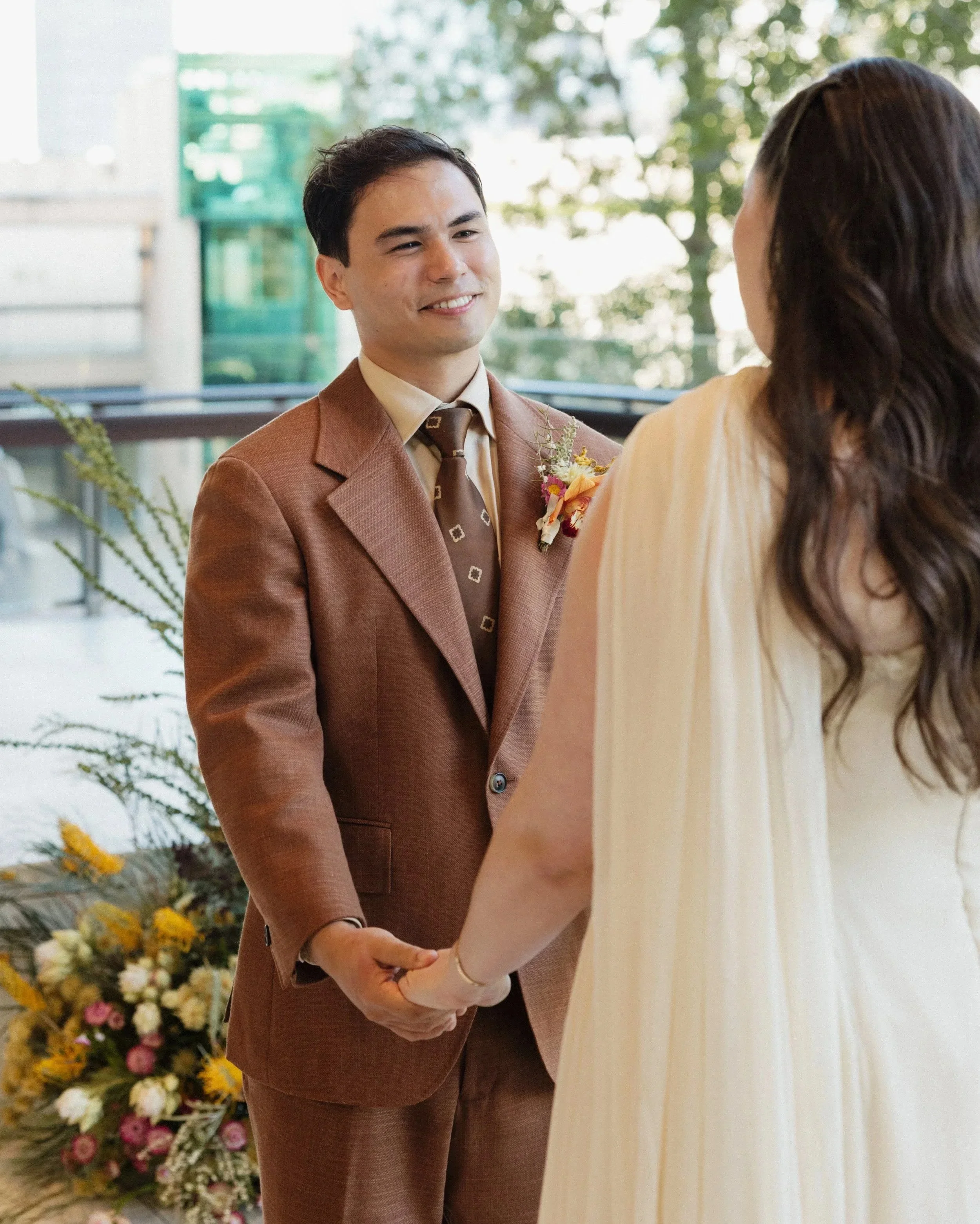 A couple holding hands during their wedding ceremony, with a beautiful floral arrangement in the background and the groom dressed in a brown Aleksandar Stately Made to measure suit and tie.