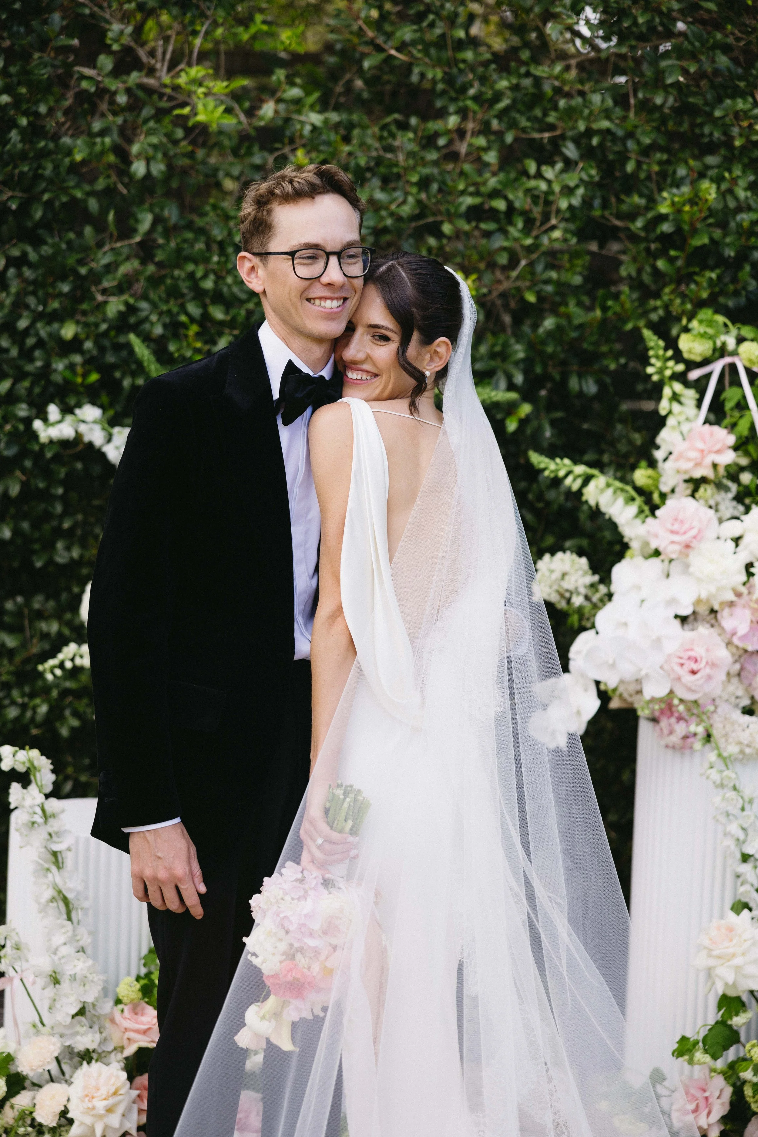 A happy couple on their wedding day, standing close to each other outdoors, groom dressed in custom tailored Alekandar Stately Suit.